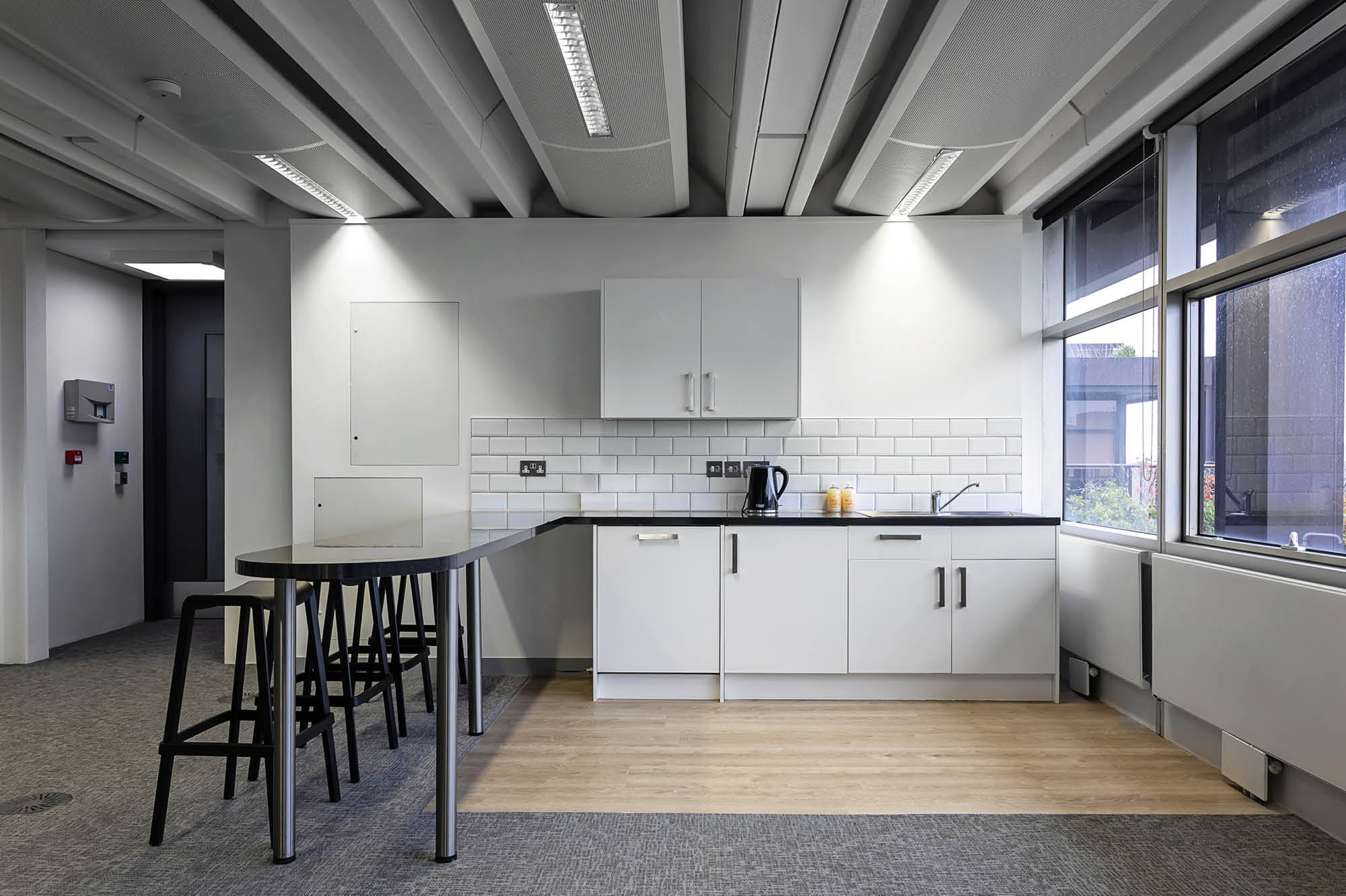 Modern office kitchenette with white cabinets and a black countertop. Features a sink, kettle, and two stools by a narrow black table.