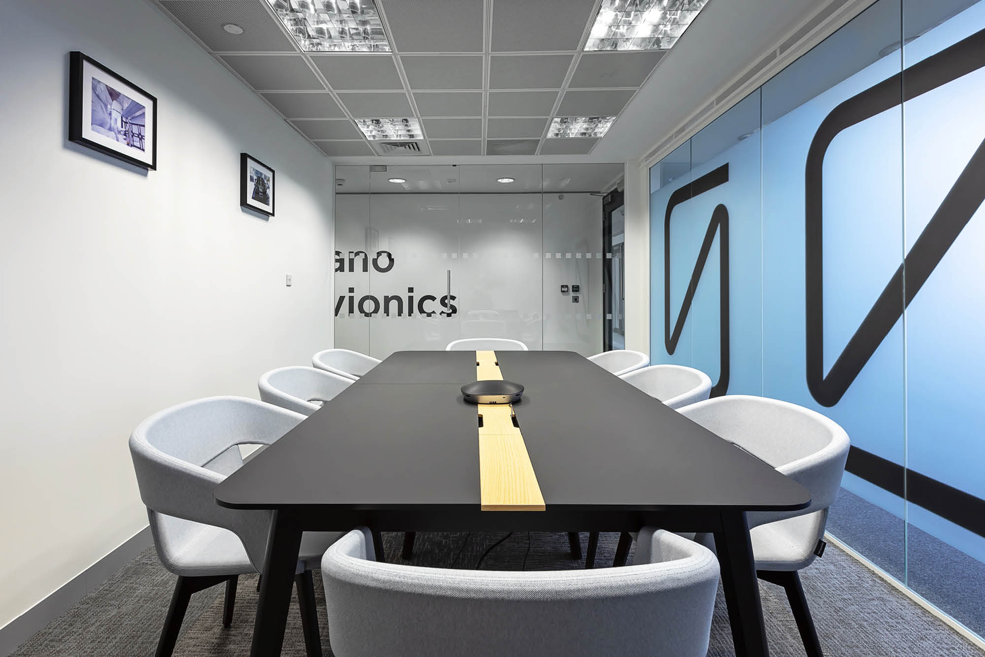 A modern conference room with a long black table surrounded by gray chairs. The room has a glass wall with blue and black geometric designs.