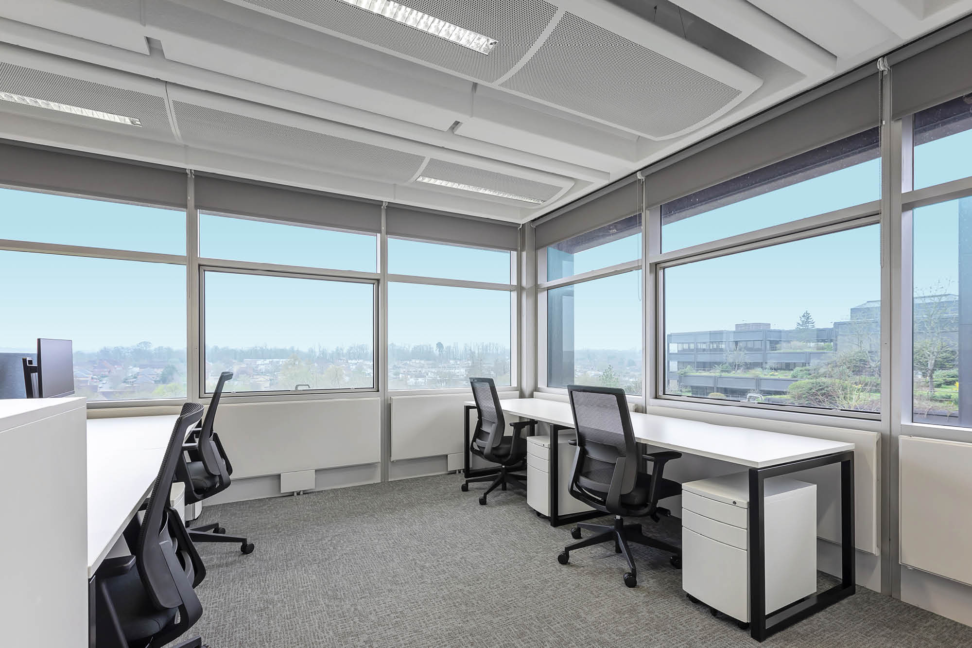 Modern office space with large windows and gray carpeting. The room has four black office chairs and three white desks, each with a drawer unit.