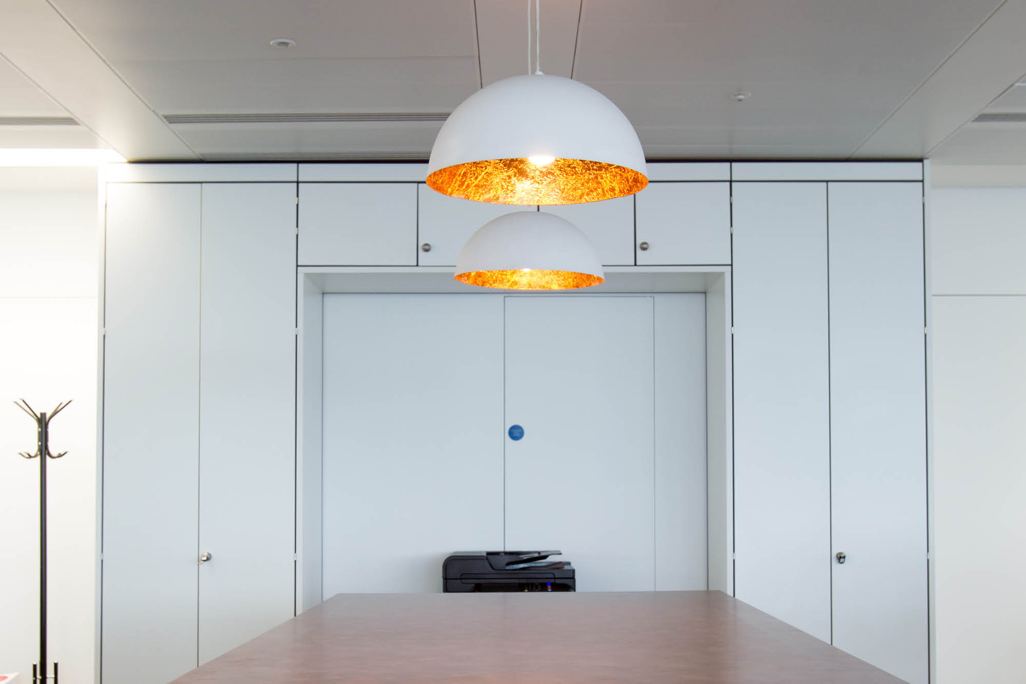 A modern office space with white cabinets and a recessed doorway. Two pendant lights with white exteriors and gold interiors hang above a long table.