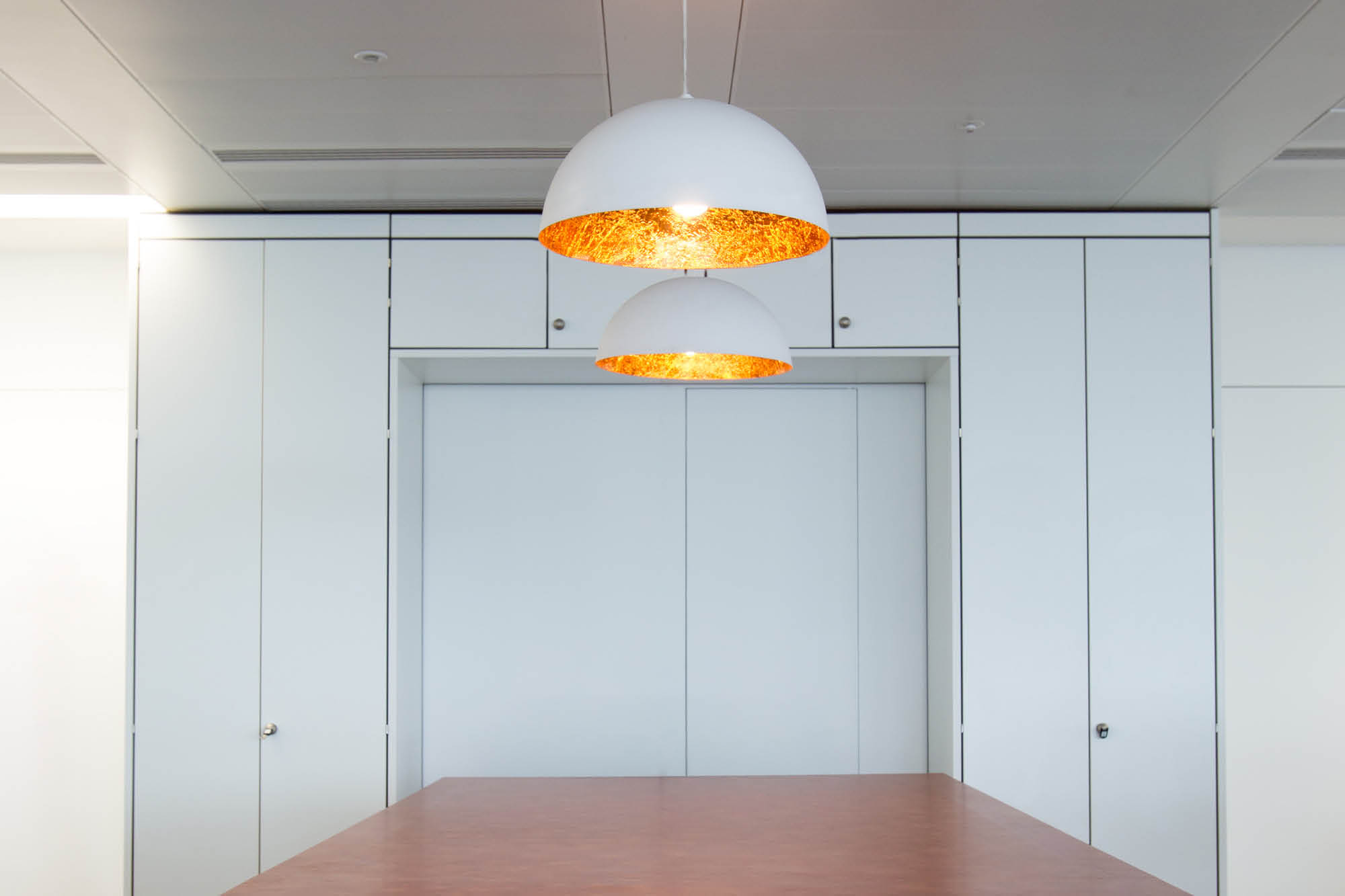 Minimalist kitchen interior with white cabinetry and two large, dome-shaped pendant lights with warm inner linings hanging.