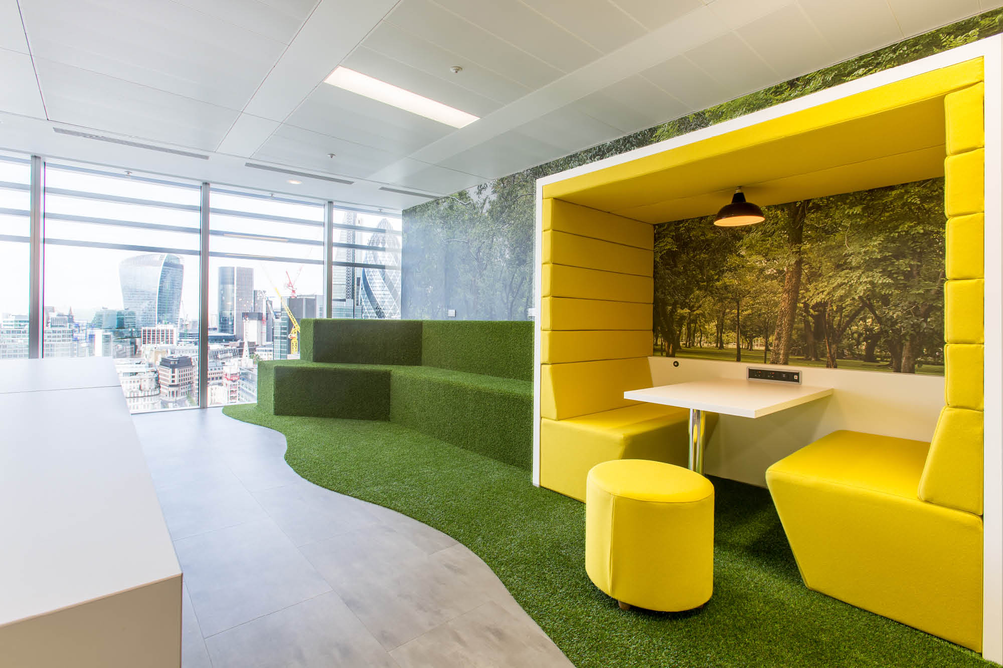 Modern office space with a bright yellow booth featuring two seats and a small table. The walls are decorated with a nature mural.