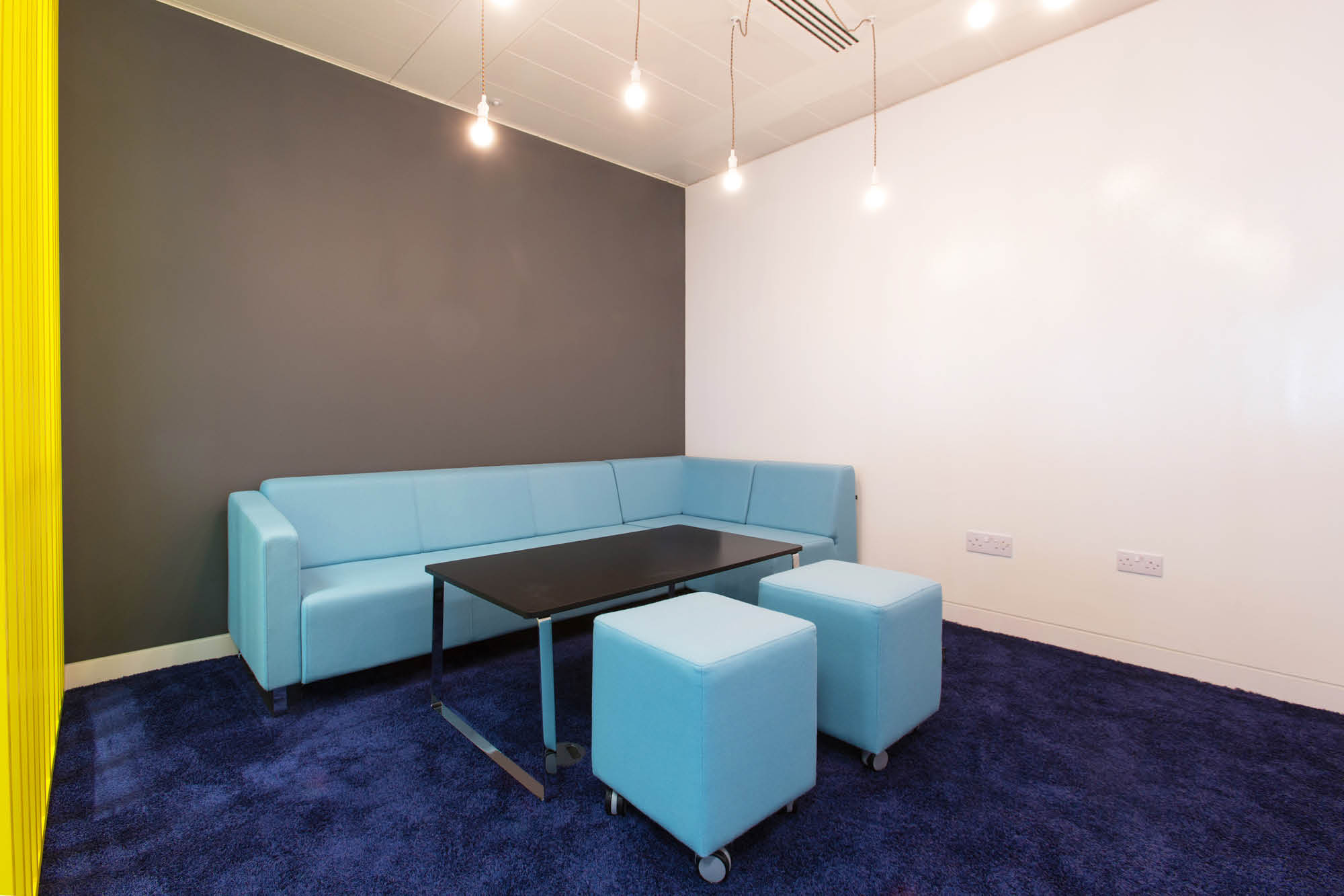 Modern office lounge with a light blue sectional sofa, two matching ottomans, and a black rectangular table.
