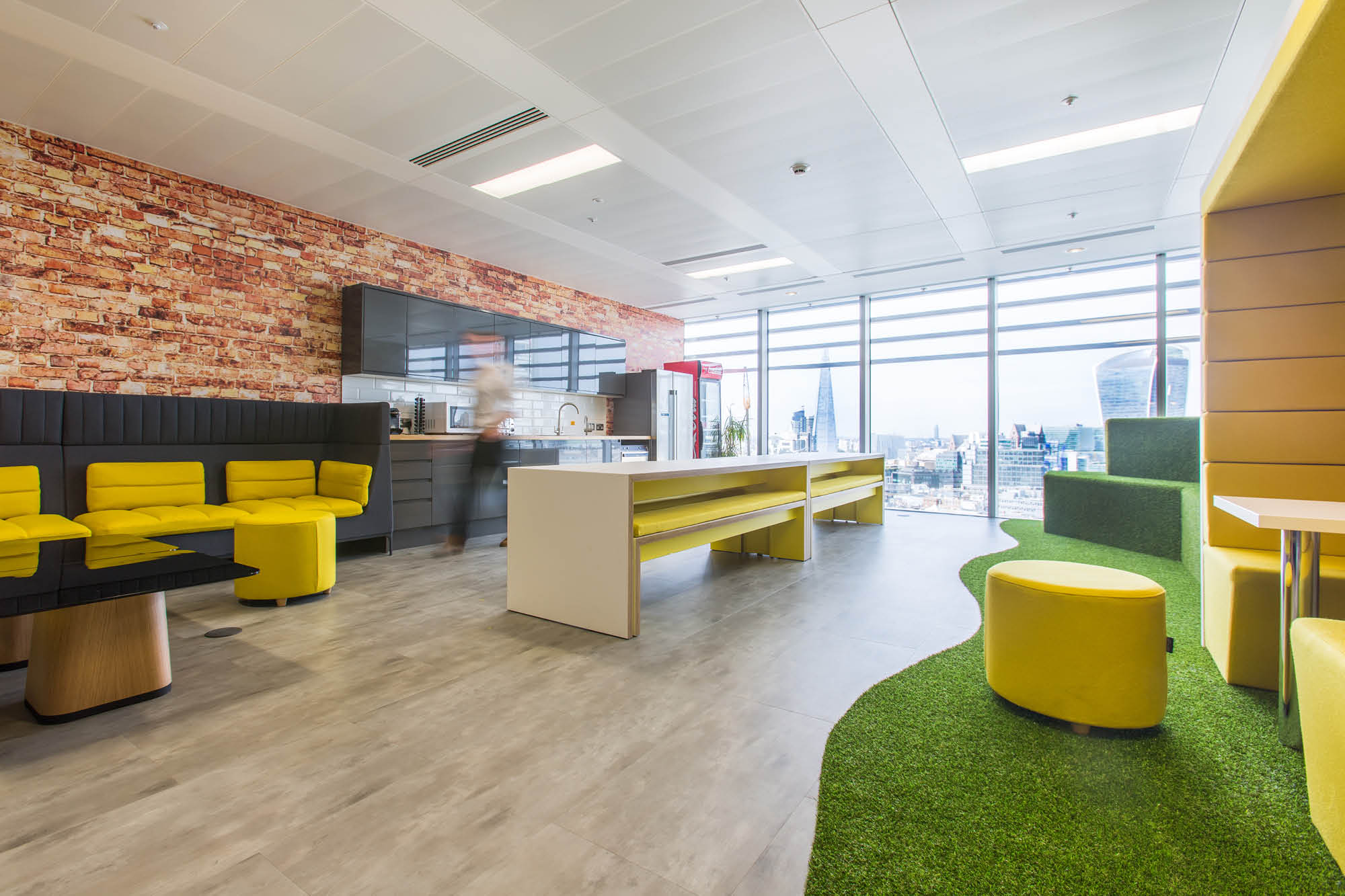 Modern office space with yellow seating, a central white table, and a faux grass area. A brick accent wall and large windows reveal a cityscape.