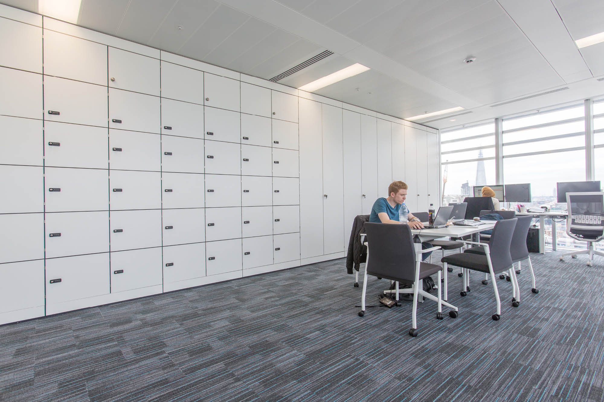 A modern office space with gray carpet and bright lighting. One person works at a table with papers and a laptop.