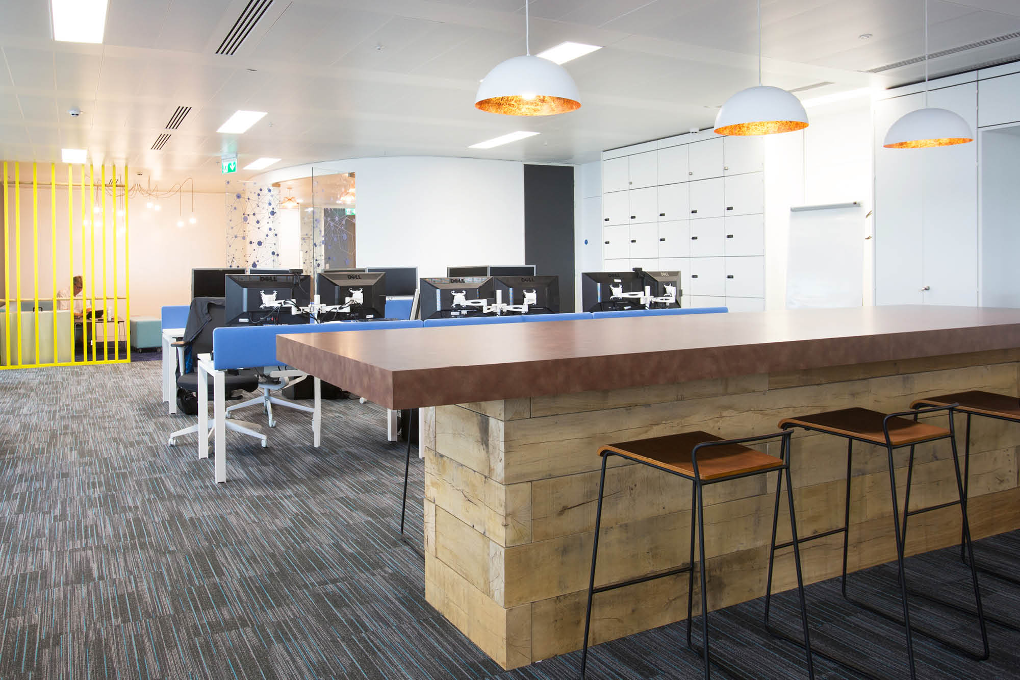 Modern office space with open-plan layout. Features a large wooden table with high stools and several computer workstations.