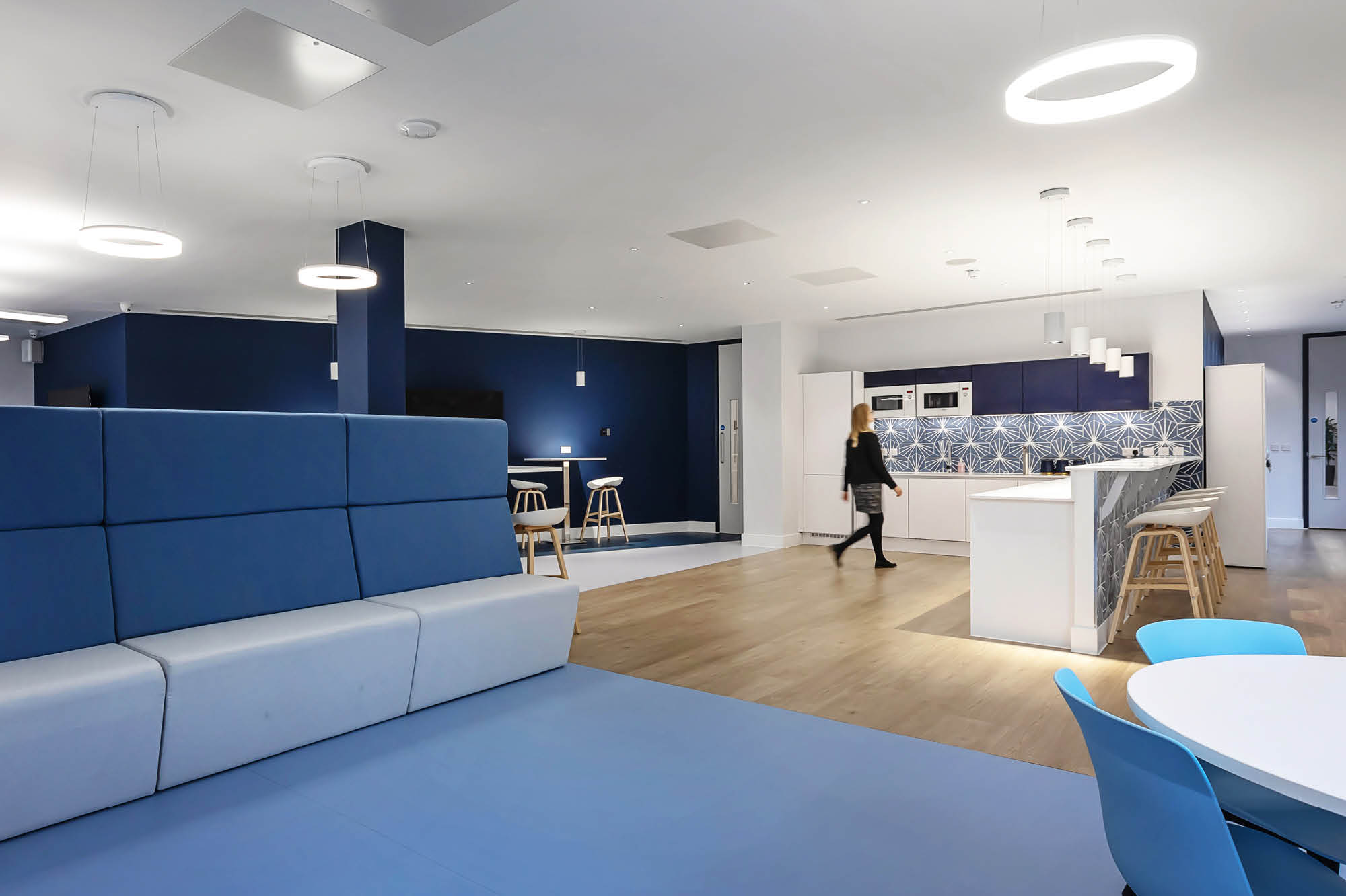 Modern office lounge with blue-and-white decor, featuring a kitchen area, circular ceiling lights, and bar stools.