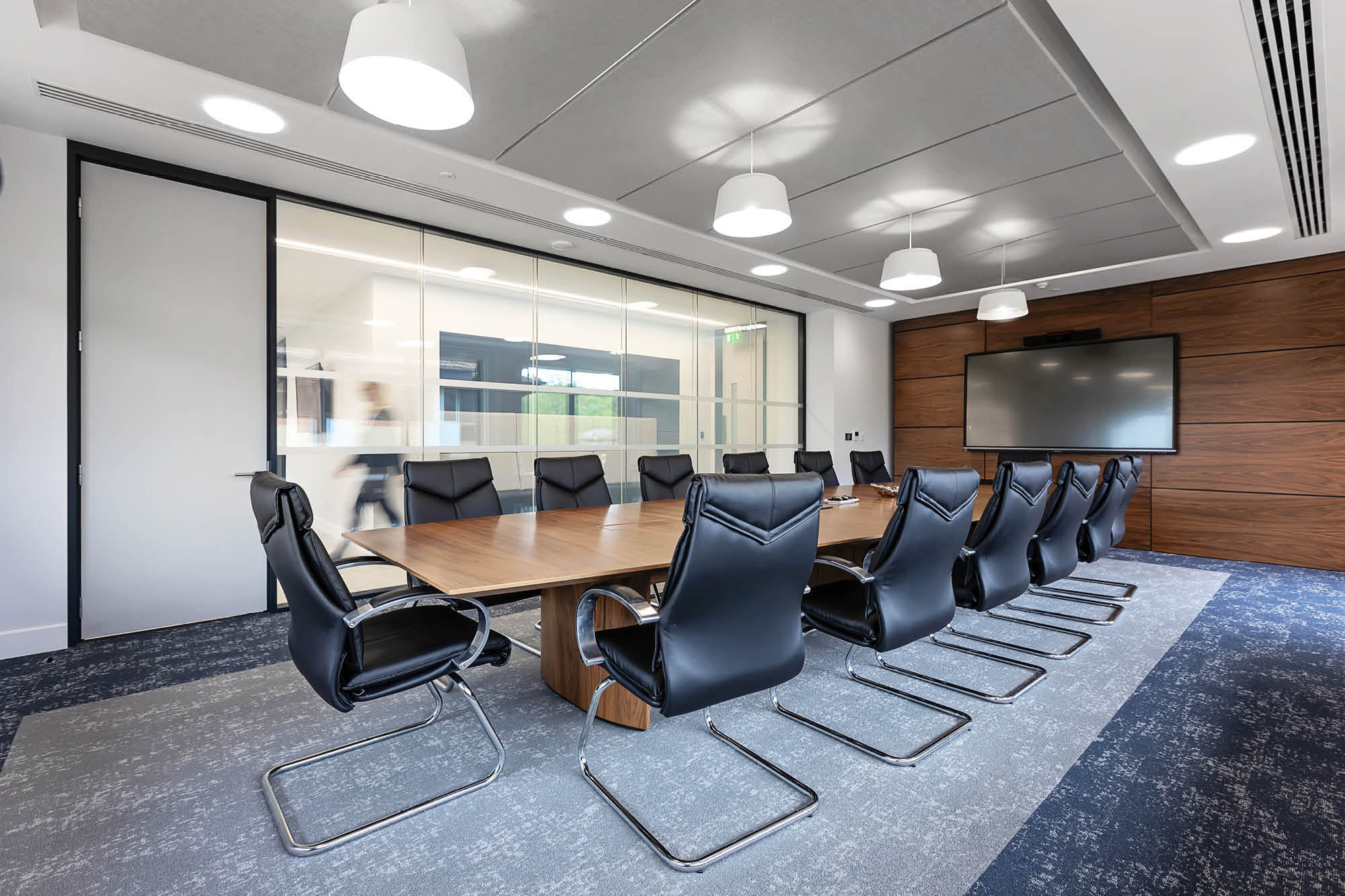 A modern conference room with a long wooden table surrounded by black leather chairs.