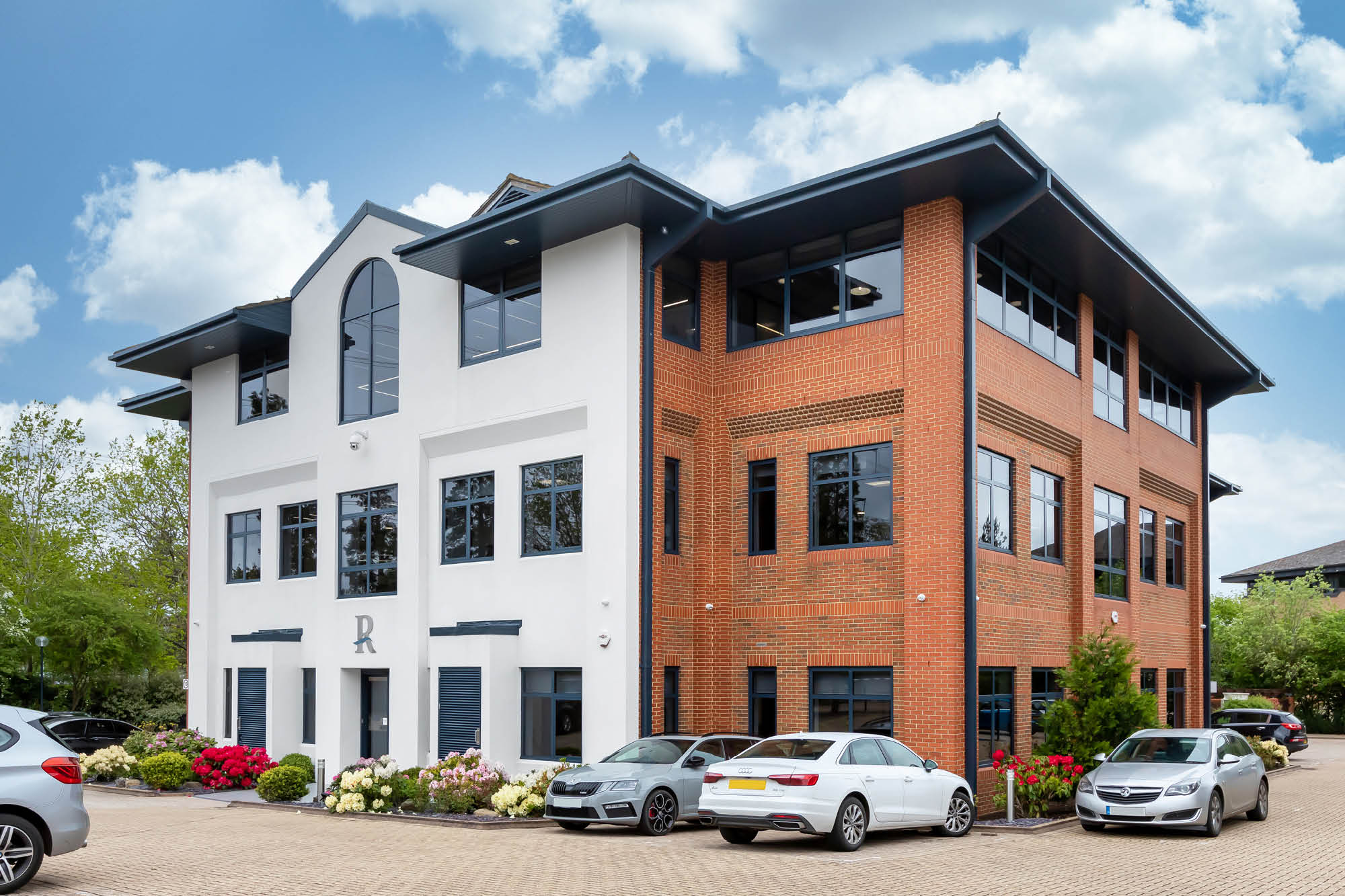 A modern, two-story office building with a mix of white and red brick facade. Several cars are parked in front on a paved surface.