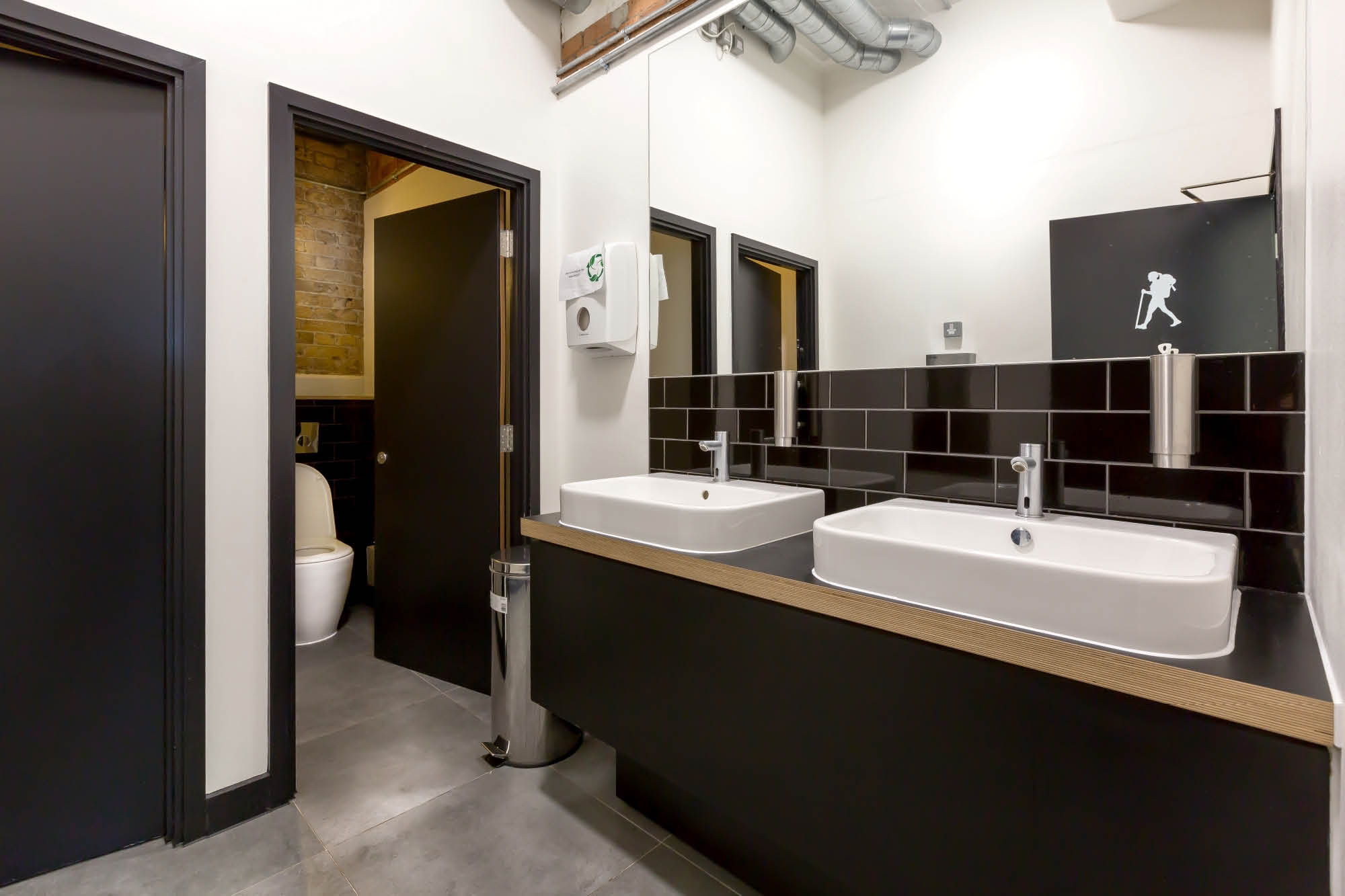 Modern restroom with two white rectangular sinks set in a black countertop, large mirror above. To the left, an open door reveals a toilet cubicle.