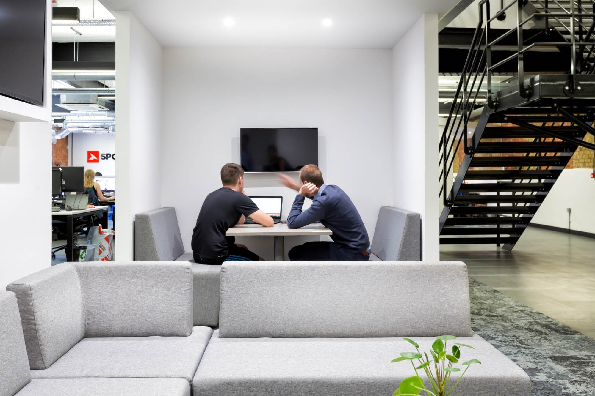 Two people sit at a table in a modern office, one gesturing towards a laptop. A TV hangs on the wall behind them.