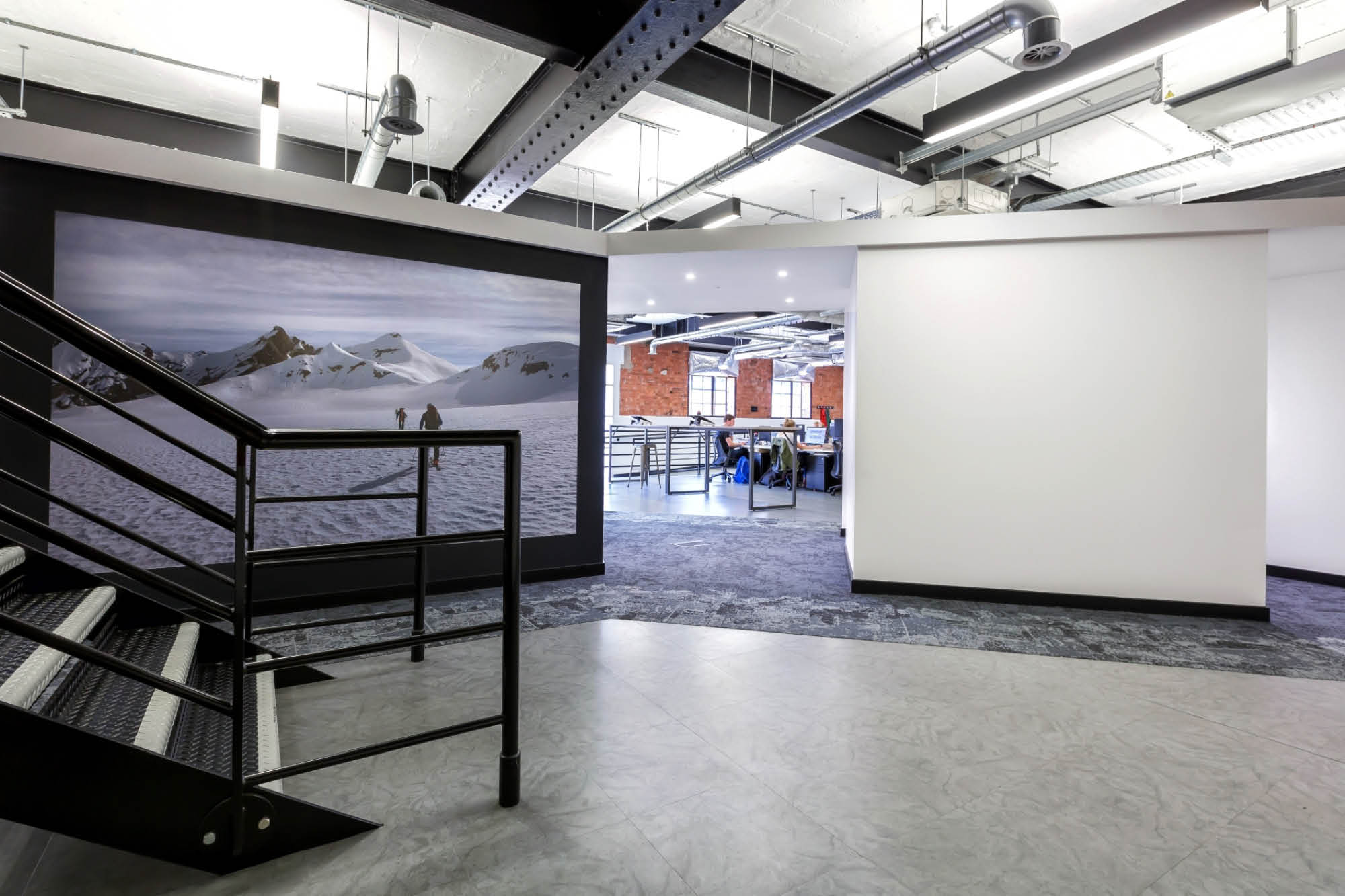 Modern office interior with glass walls, exposed ceilings, and a metal staircase. A large photograph of a snowy mountain landscape is mounted.