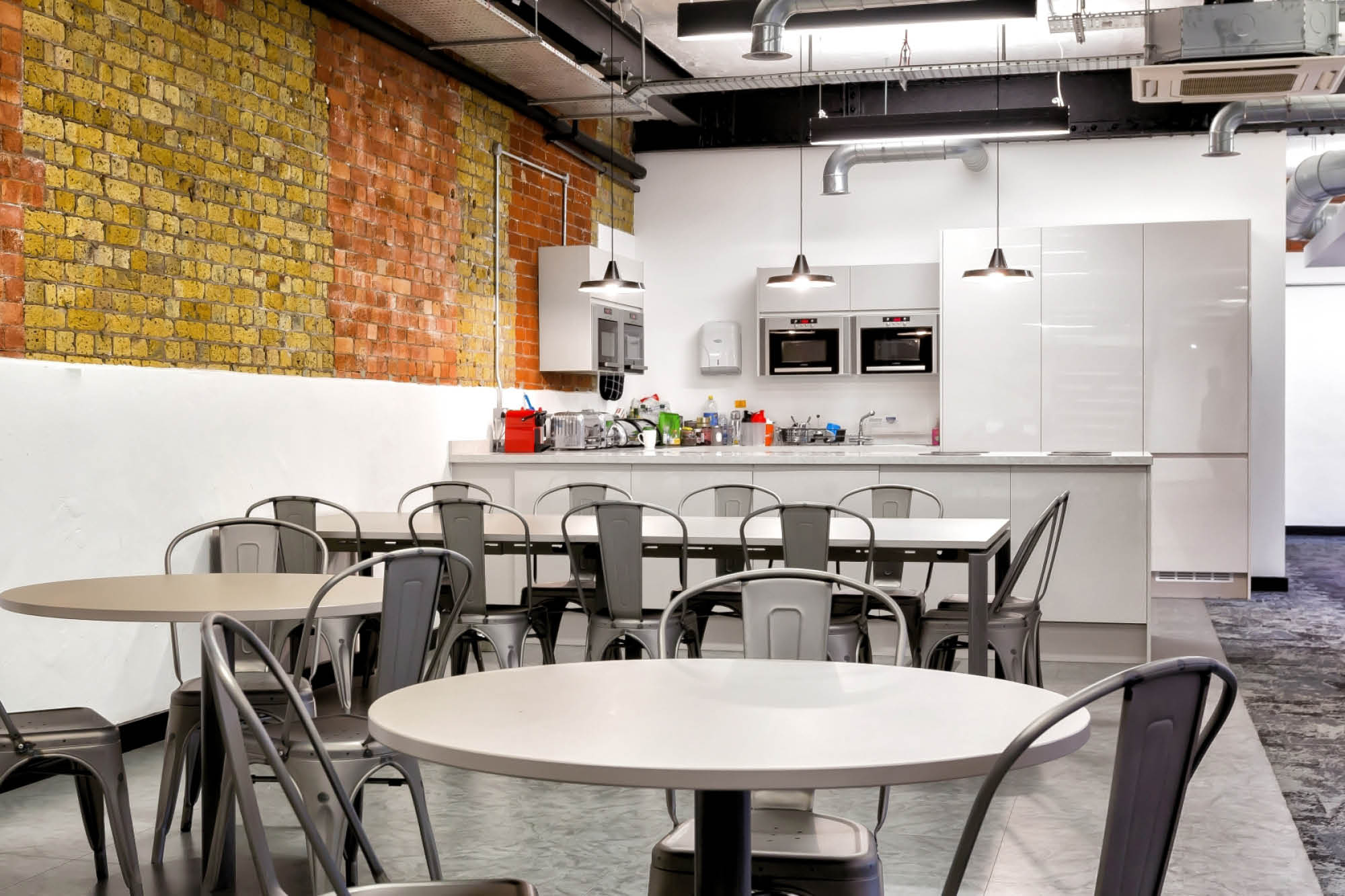 Modern office kitchen with exposed brick wall, round tables, and metal chairs. A countertop with coffee machines and a sink.