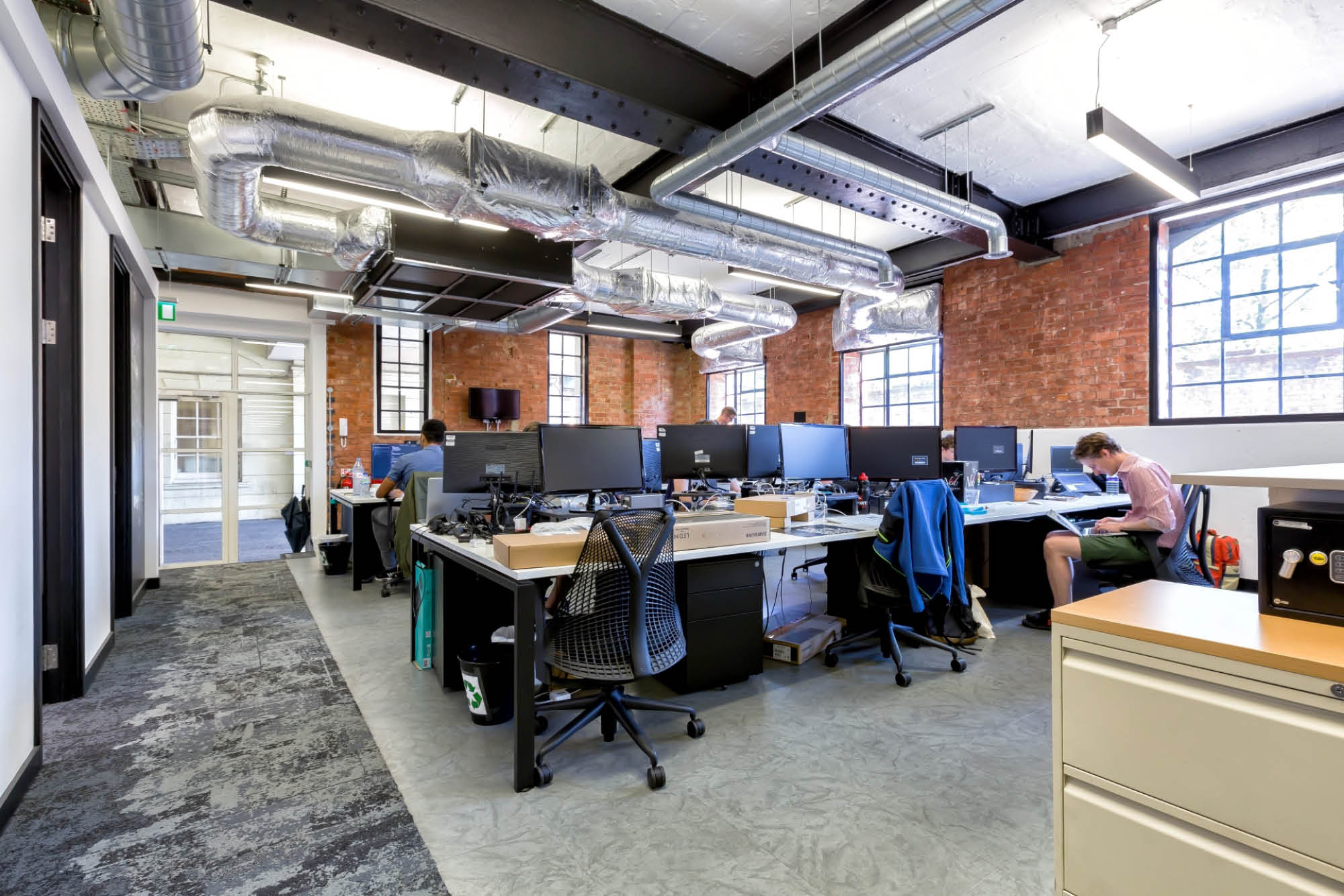 A modern open office space with high ceilings, exposed brick walls, and visible ductwork. Several desks with computers are occupied.