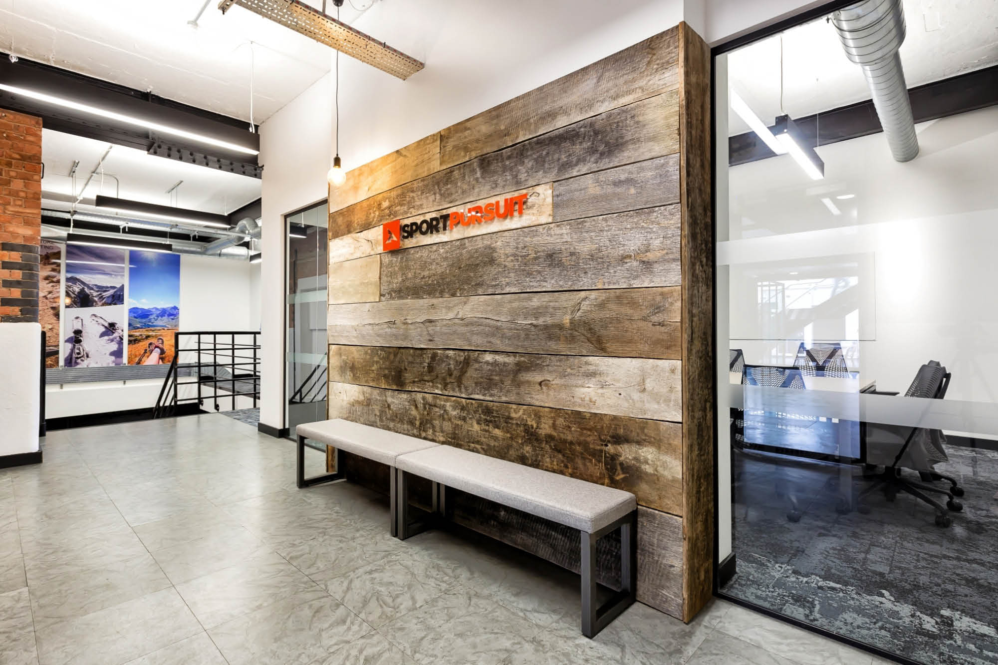 Modern office hallway with a rustic wooden wall featuring a logo. A bench is placed against the wall. Glass doors lead to a conference room.