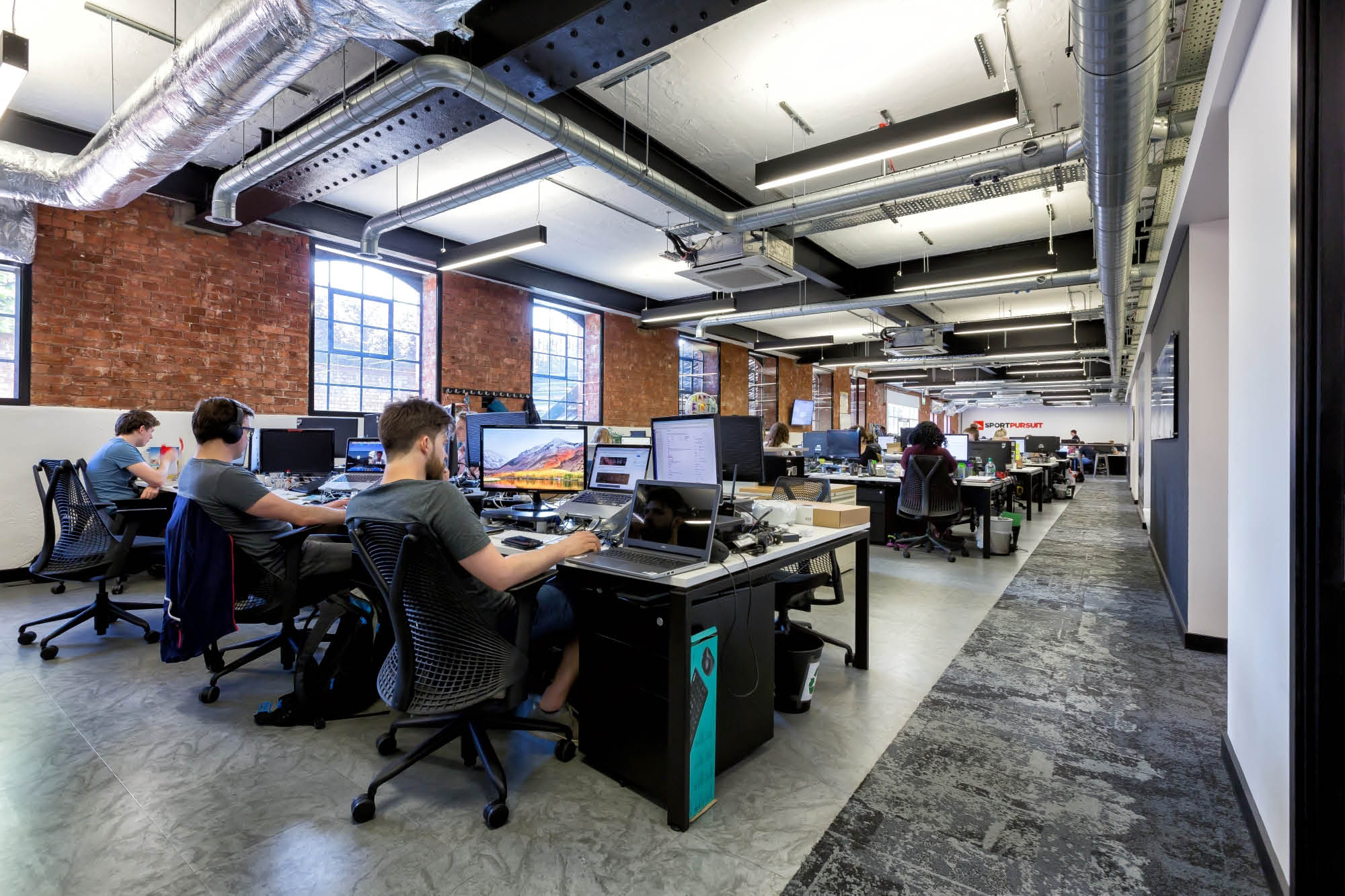 An open office space with exposed brick walls and ductwork. Several people are working at desks with computers.