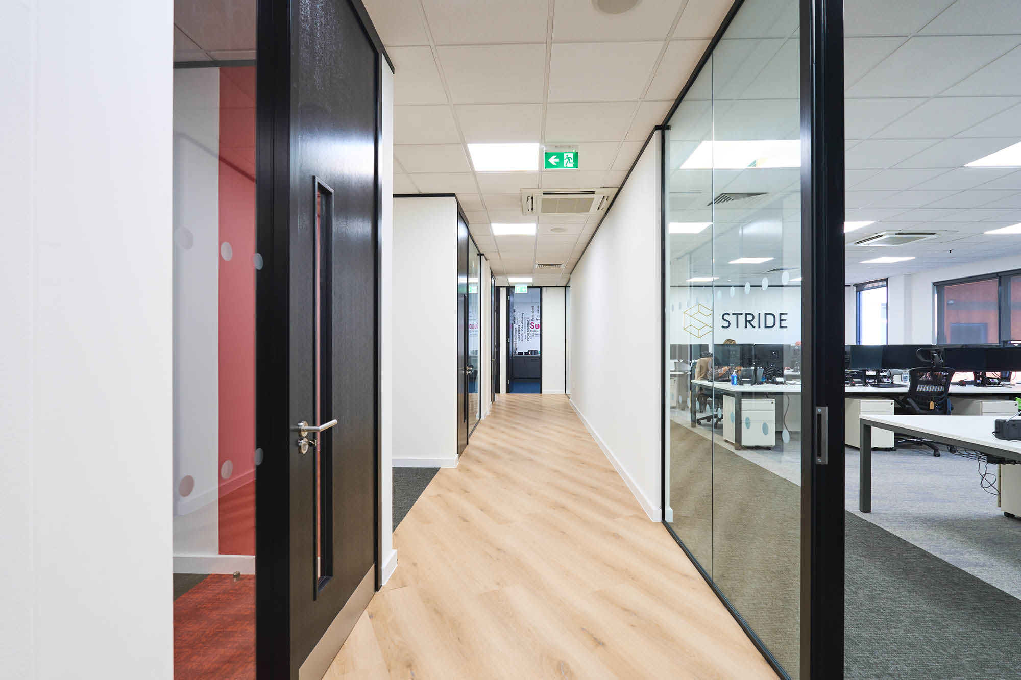 A modern office hallway with wooden flooring and glass-walled sections on both sides. On the right, desks with computers.