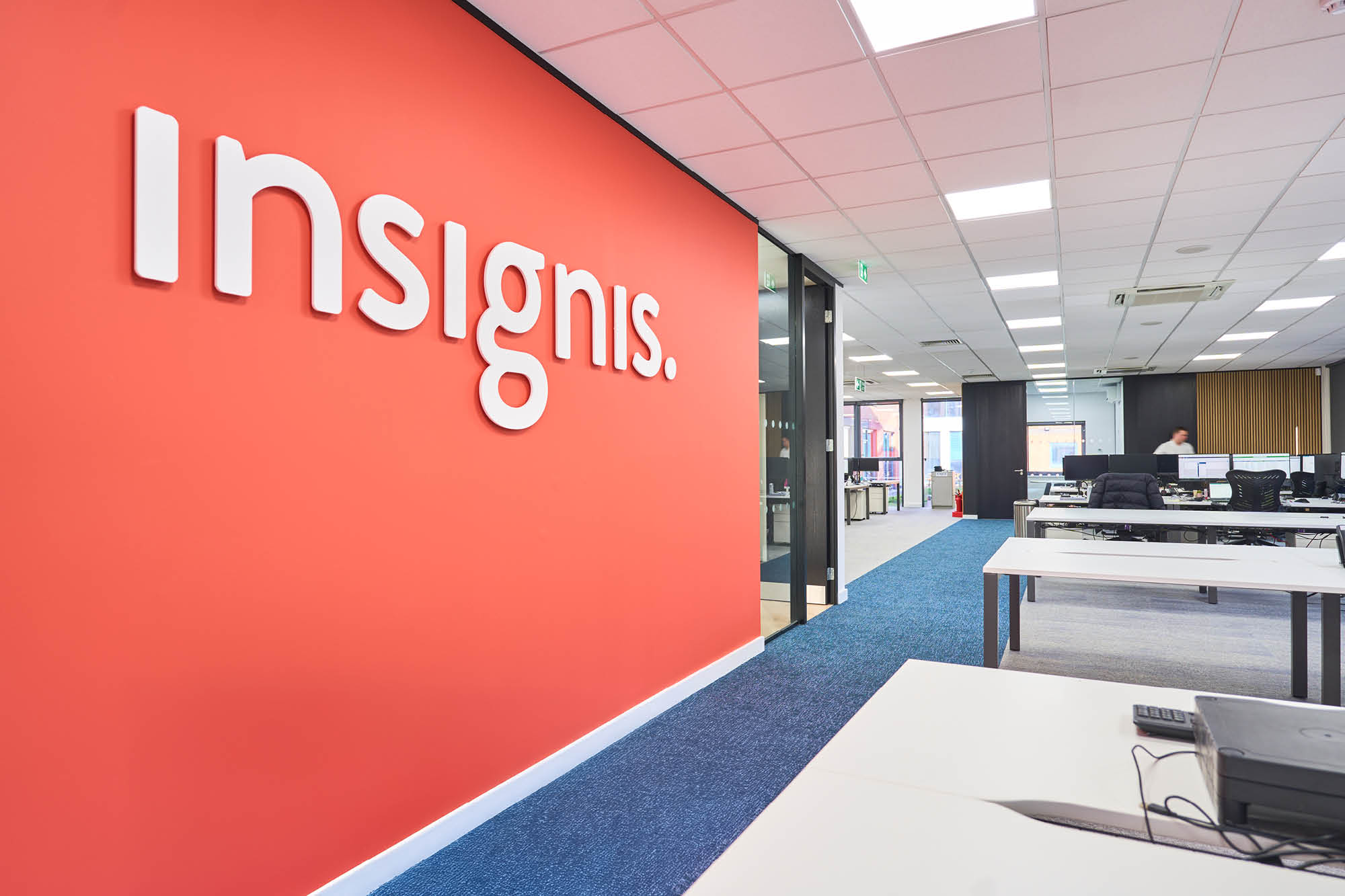 Modern office space with a red wall displaying the word Insignis in white letters. The room features blue carpet, white desks with computers.