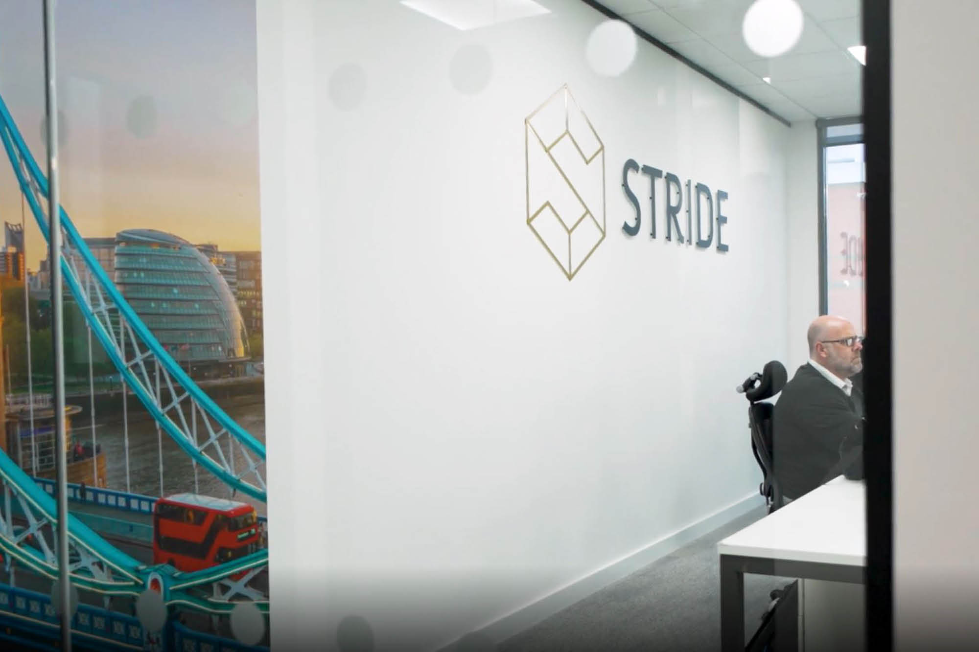 Man seated at a desk in a modern office with a glass wall displaying a logo that reads STRIDE. A large mural of London.