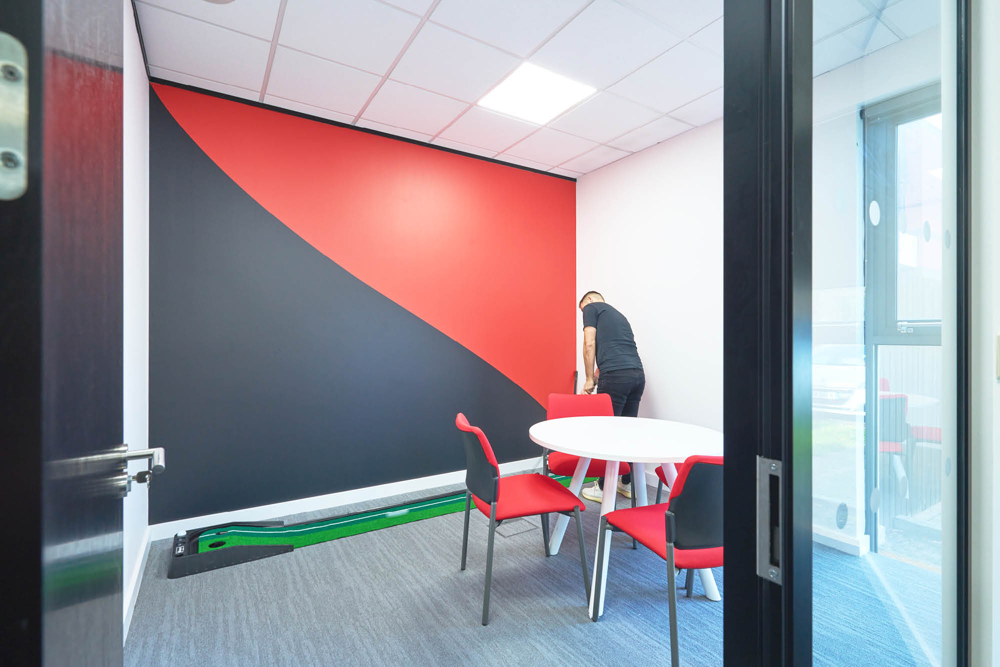 A modern office room with two red and gray chairs around a round white table. A person is adjusting a third chair near a wall with a bold red and black design.