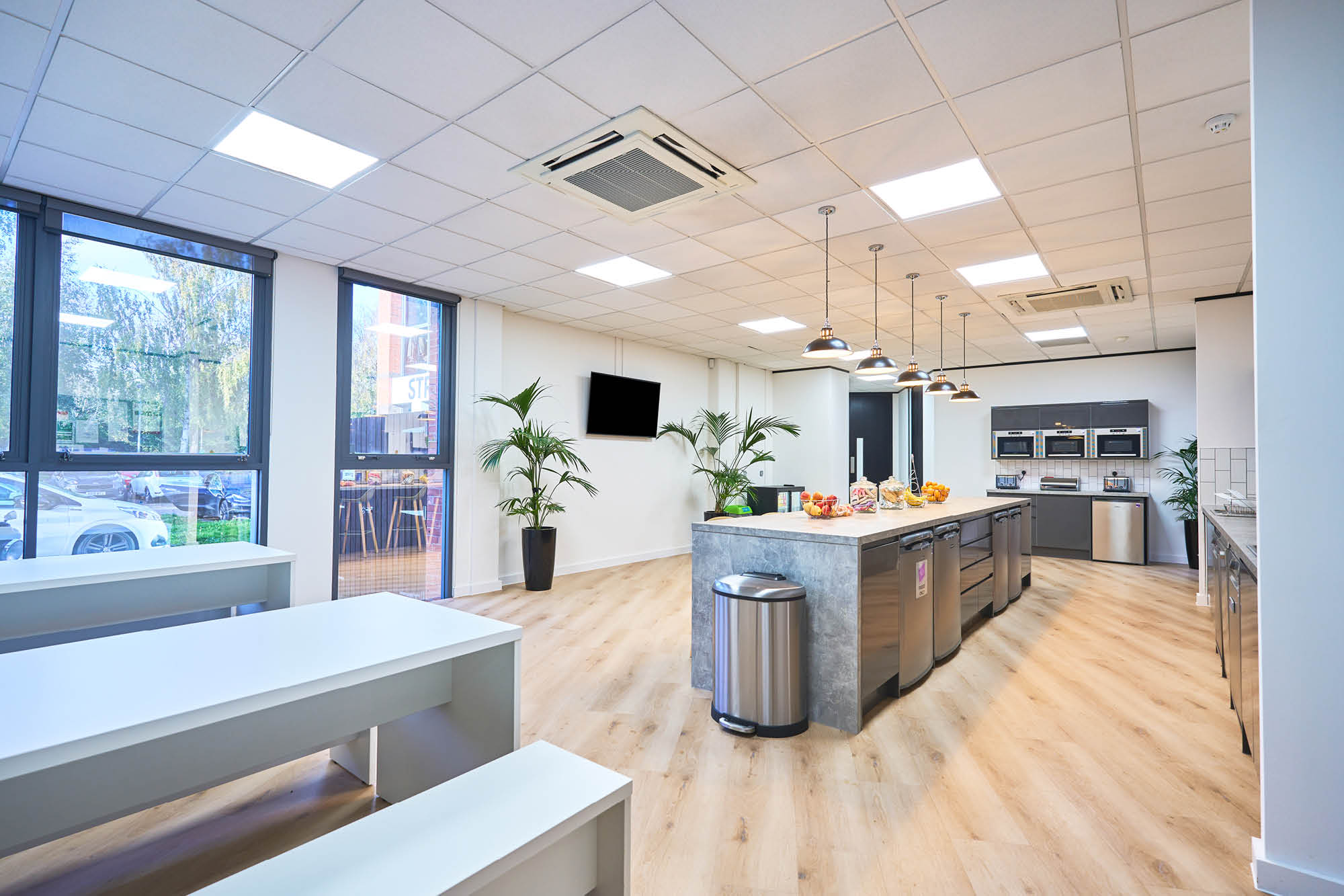 Spacious modern kitchen area with a central island, pendant lights, and light wood flooring. Large windows provide natural light.