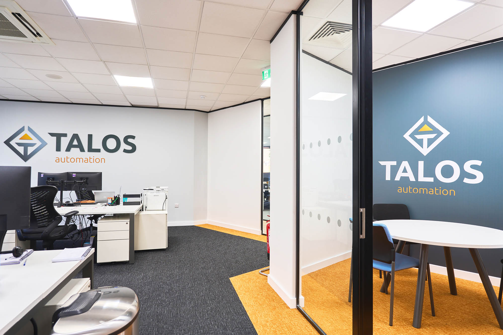Modern office interior with TALOS automation logos on the walls. The space features a desk with a computer, chairs, a small table.