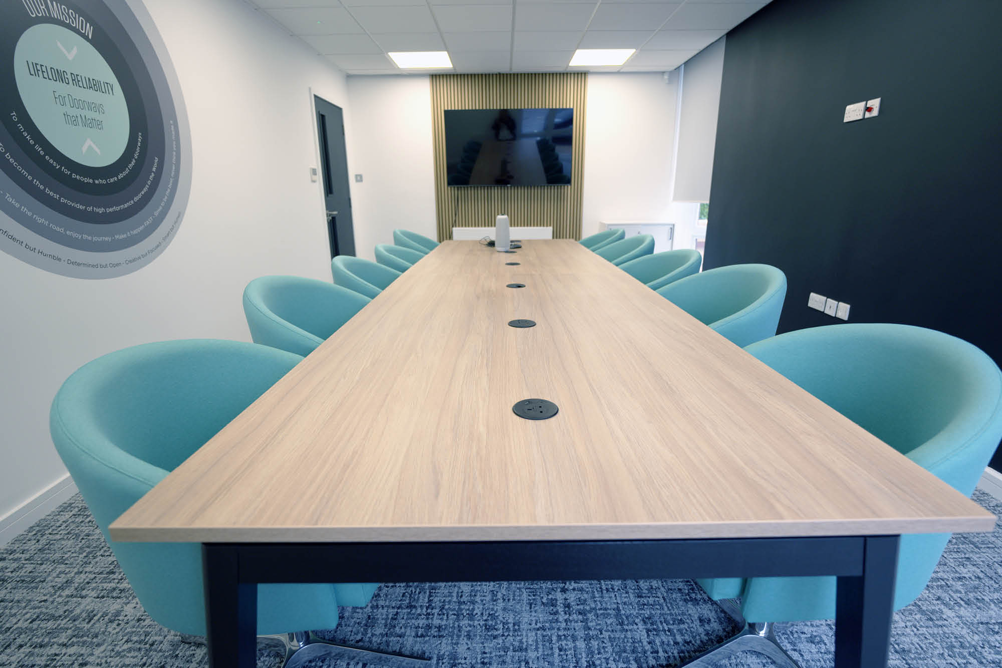 A modern conference room with a long wooden table surrounded by teal chairs. A large screen is mounted on the wall at the far end.