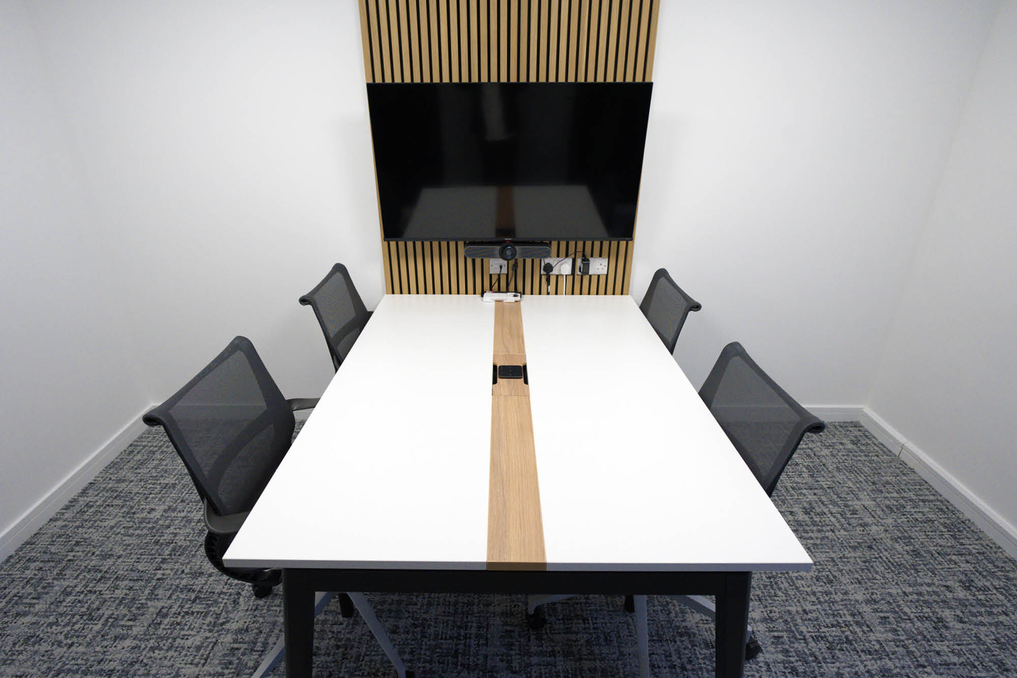 A small conference room with a modern design, featuring a long white table, four mesh chairs, and a large flat-screen TV.