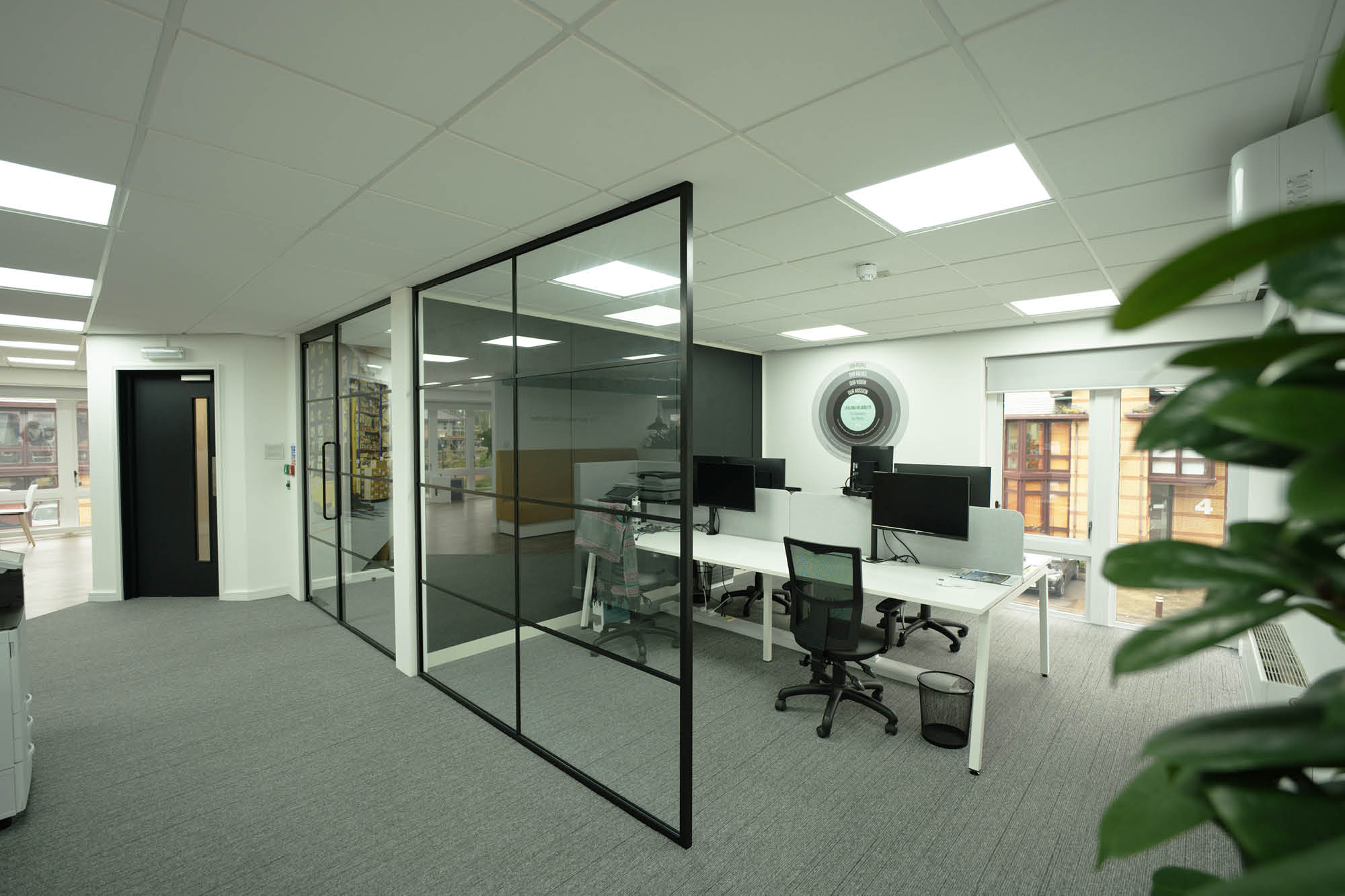 Modern office with several workstations, each equipped with computers and chairs. A glass partition separates the area.