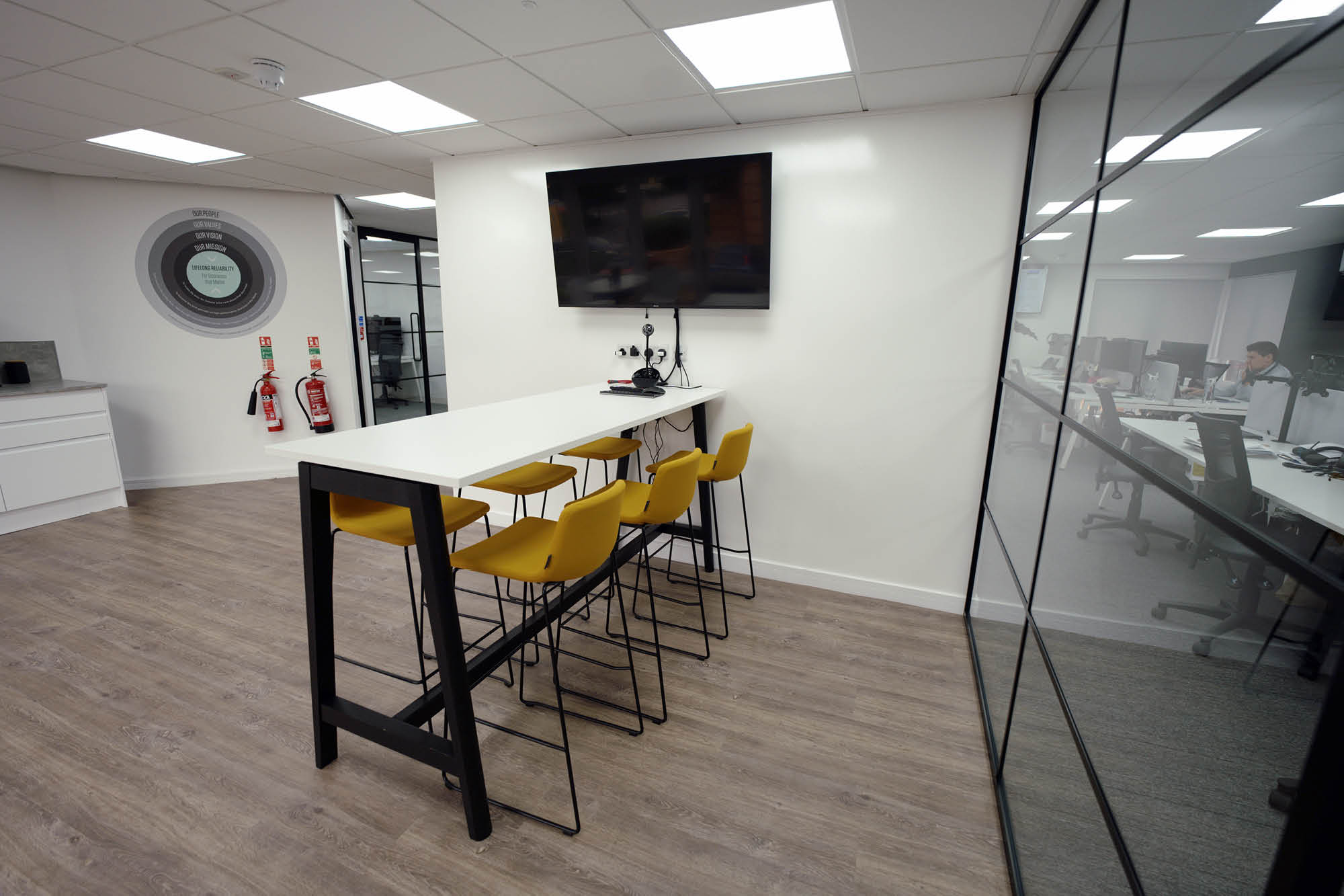 Modern office break area with a high white table and four yellow chairs. A TV is mounted on the white wall above.