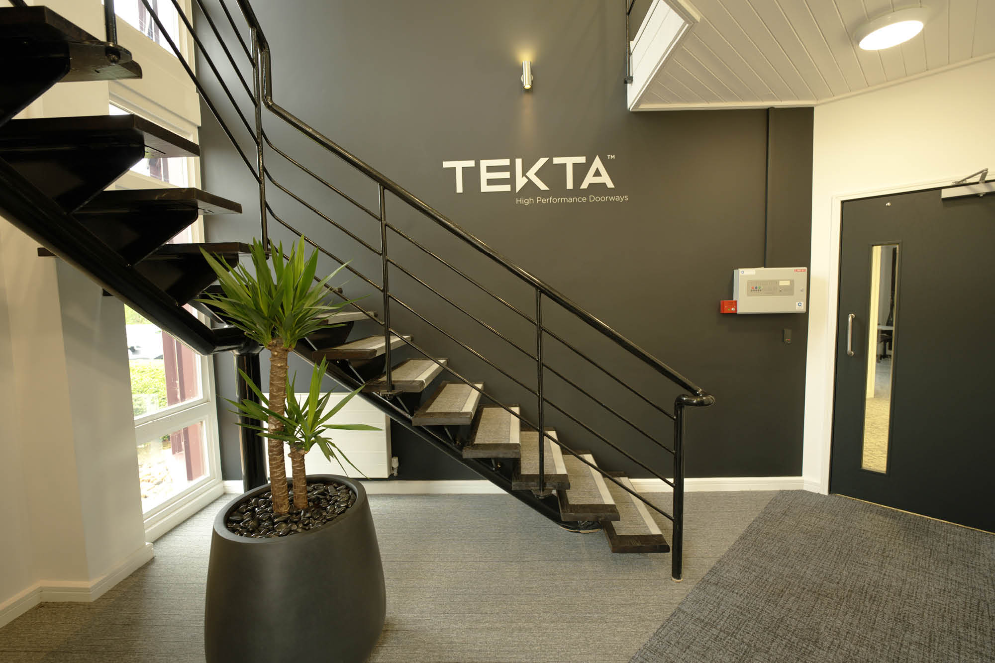 Staircase with black railings leading to the upper floor next to a potted plant. A gray wall features the text TEKTA High Performance Doorways.