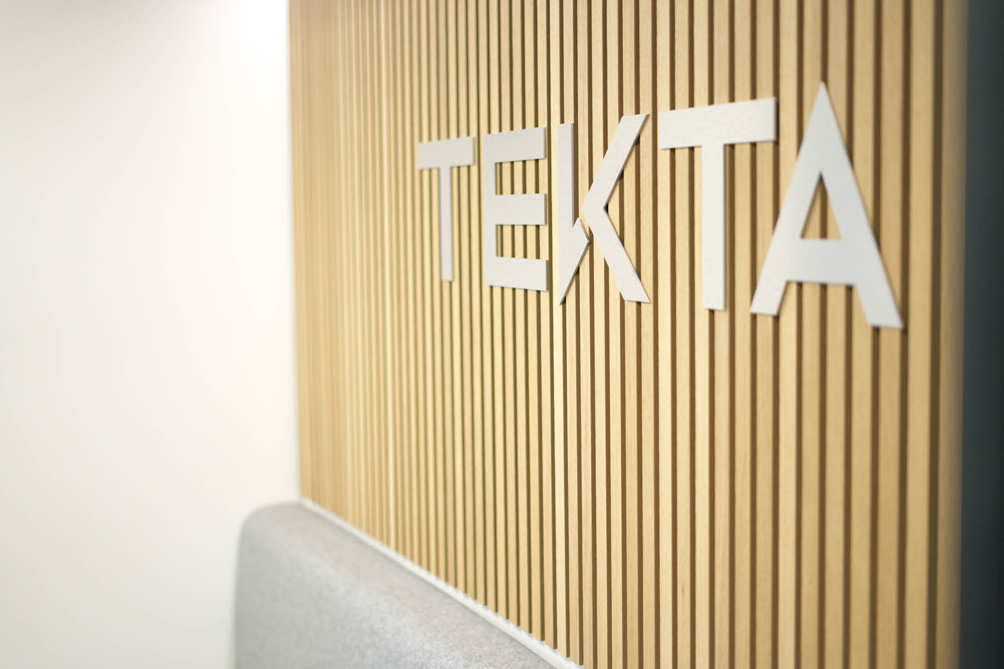 Wooden wall with vertical slats displaying the text TEKTA in white, next to a light gray upholstered bench.