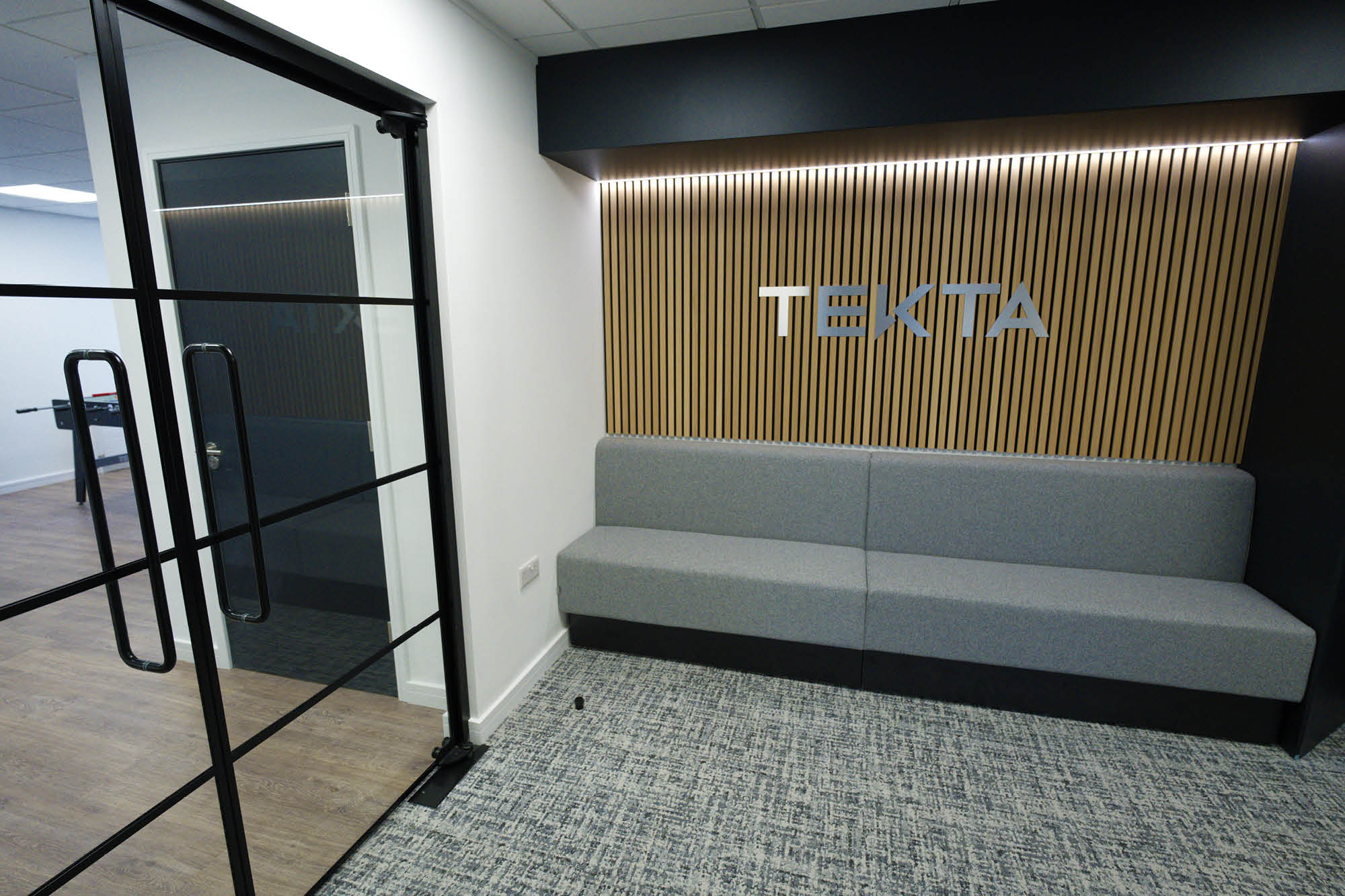 Office lobby with a gray cushioned bench against a wooden slat wall featuring the word TEKTA in metallic letters.