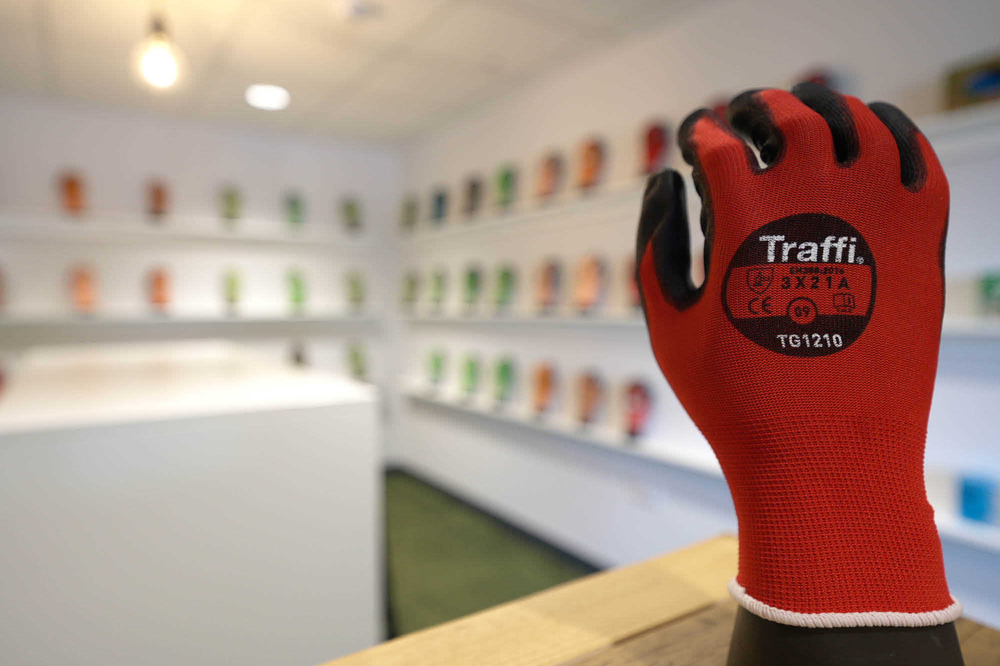 A showroom displays a variety of colorful gloves on shelves in the background. In the foreground, a red and black safety glove is mounted.