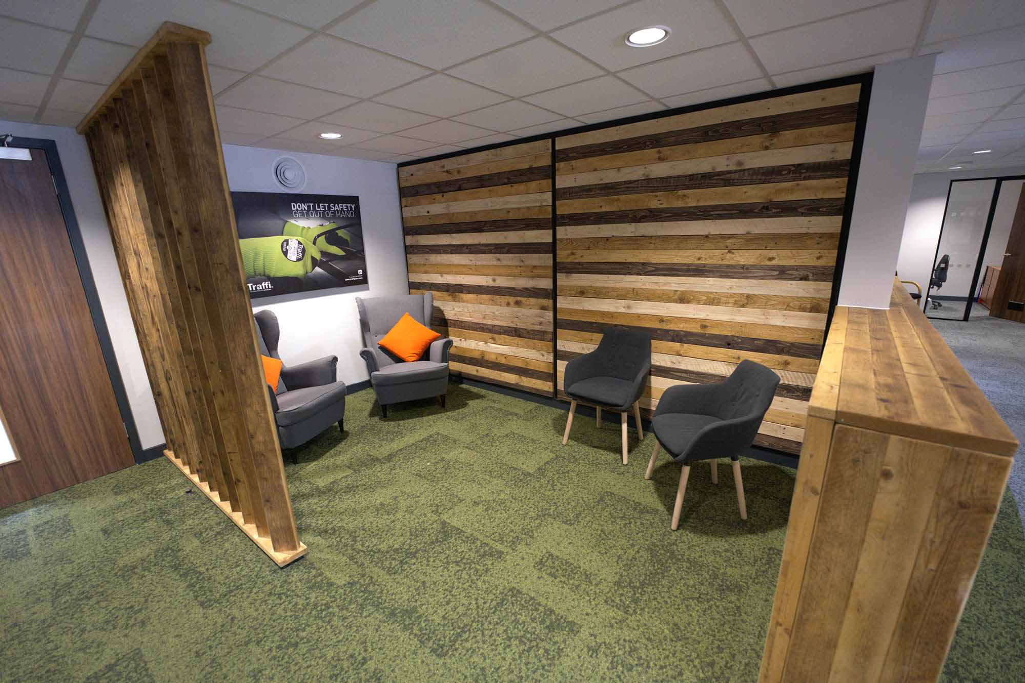 Small office lounge with green carpet, wooden wall panels, and four grey chairs, two of which have orange cushions.