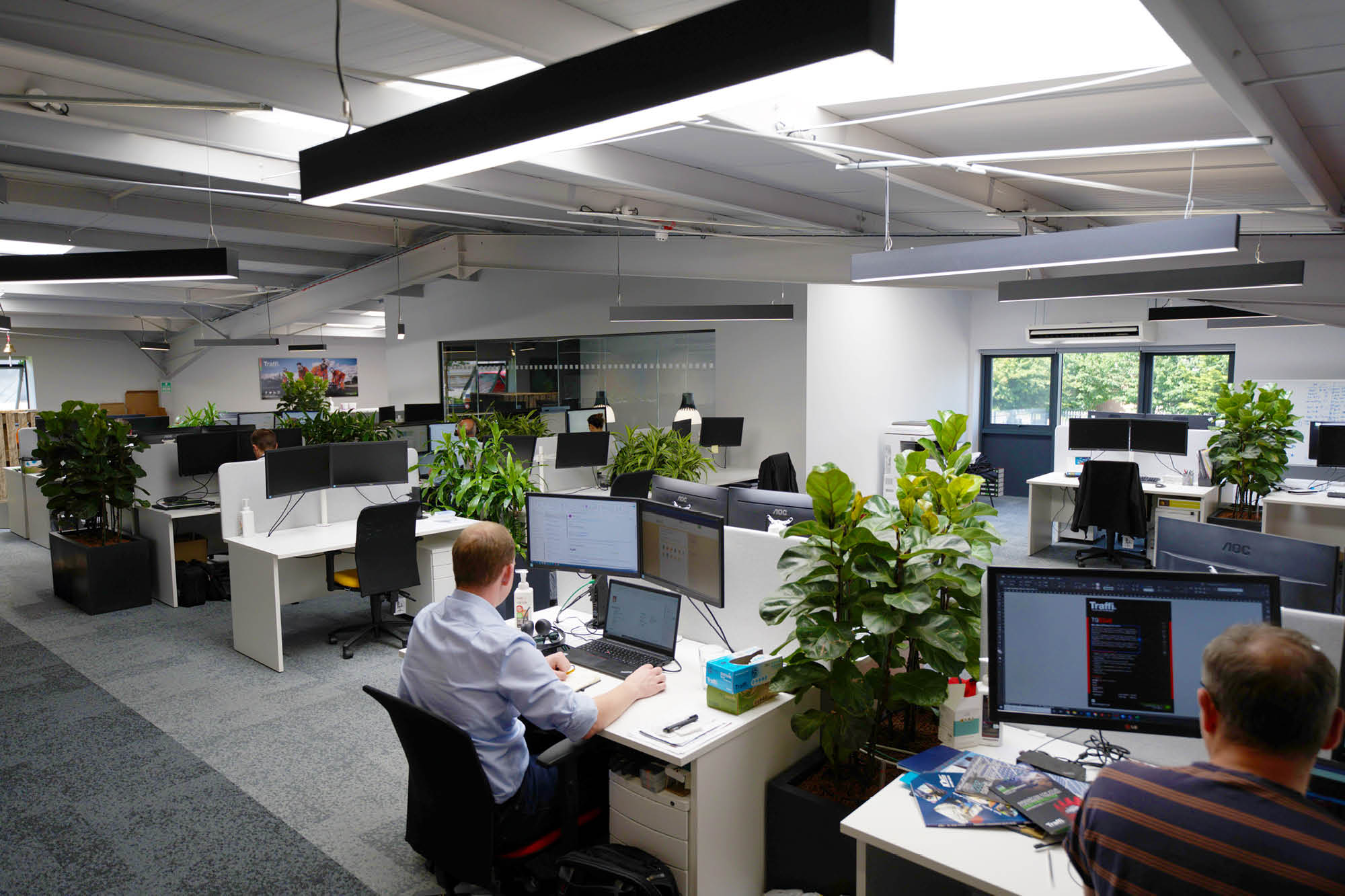 Open-plan office with several people working at desks with computers. The space has overhead lighting.