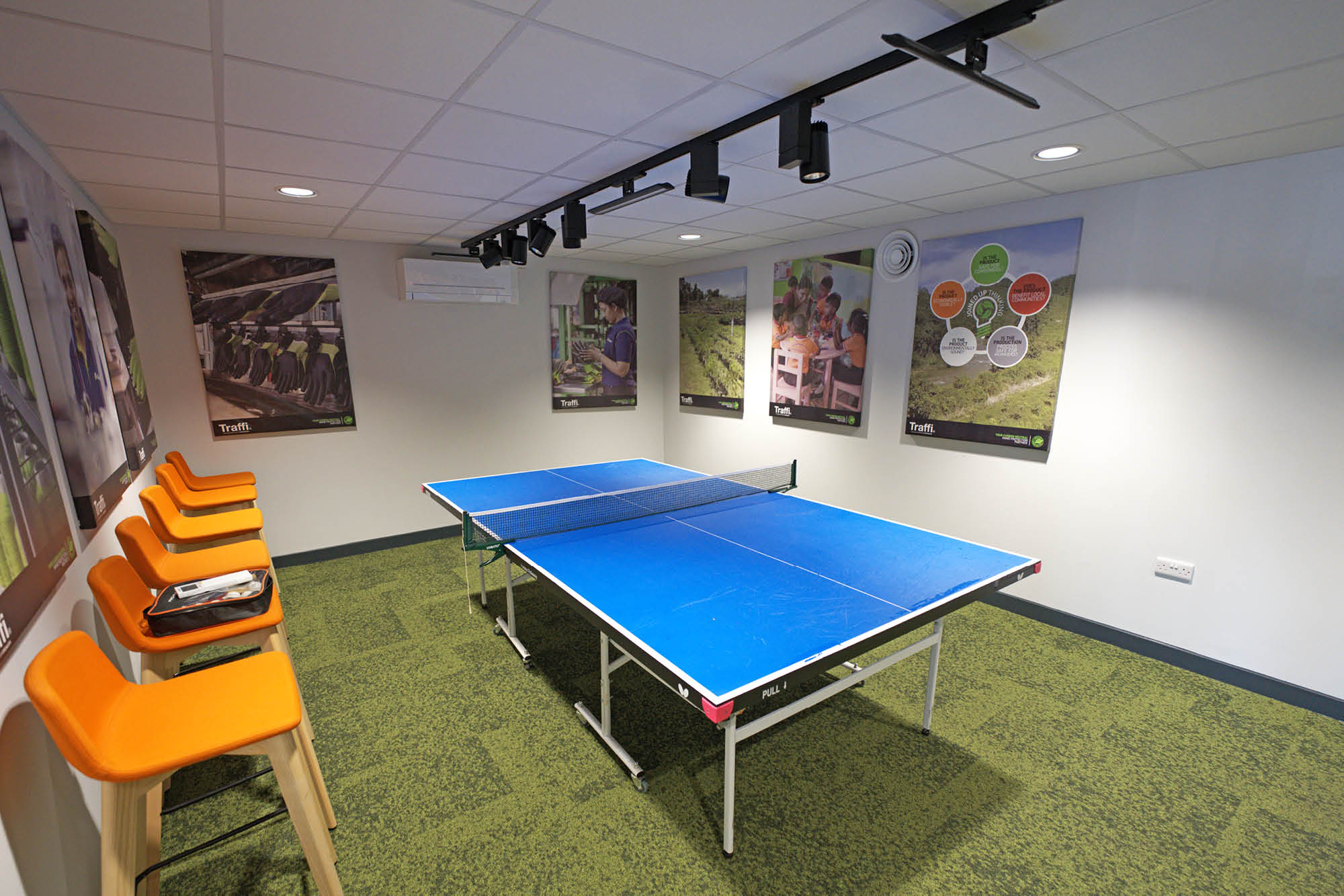 Room with a ping pong table, surrounded by orange high stools along one wall. The floor is carpeted in green, and there are various posters on the walls.