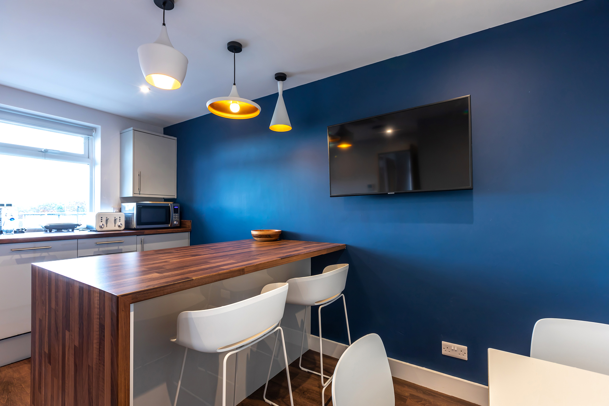 Modern kitchen with blue accent wall, three pendant lights, and a wall-mounted TV. A wooden island features four white bar stools.