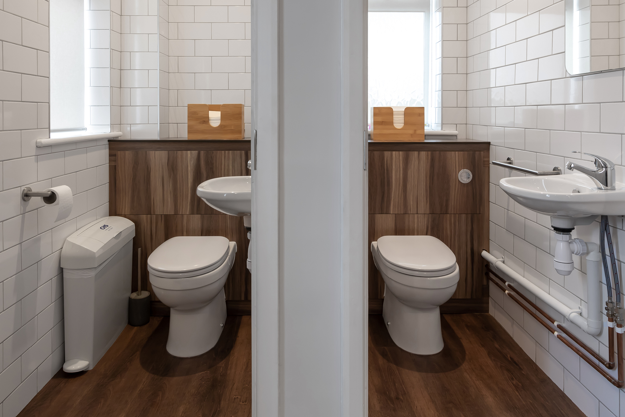 Two adjacent bathrooms separated by a thin wall, each with a white toilet, a wall-mounted sink, and a brown wood cabinet.