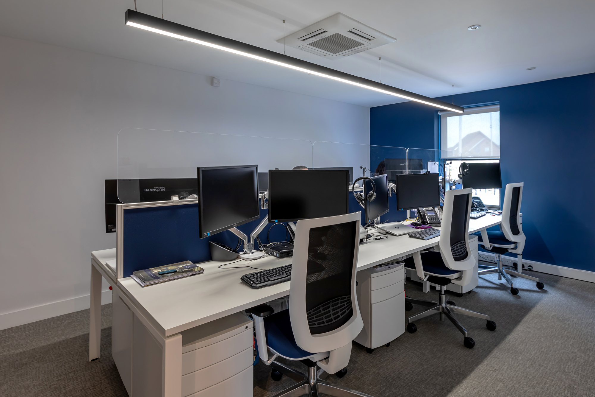 A modern open-plan office with a row of four workstations. Each has a chair, computer monitor, and office supplies.