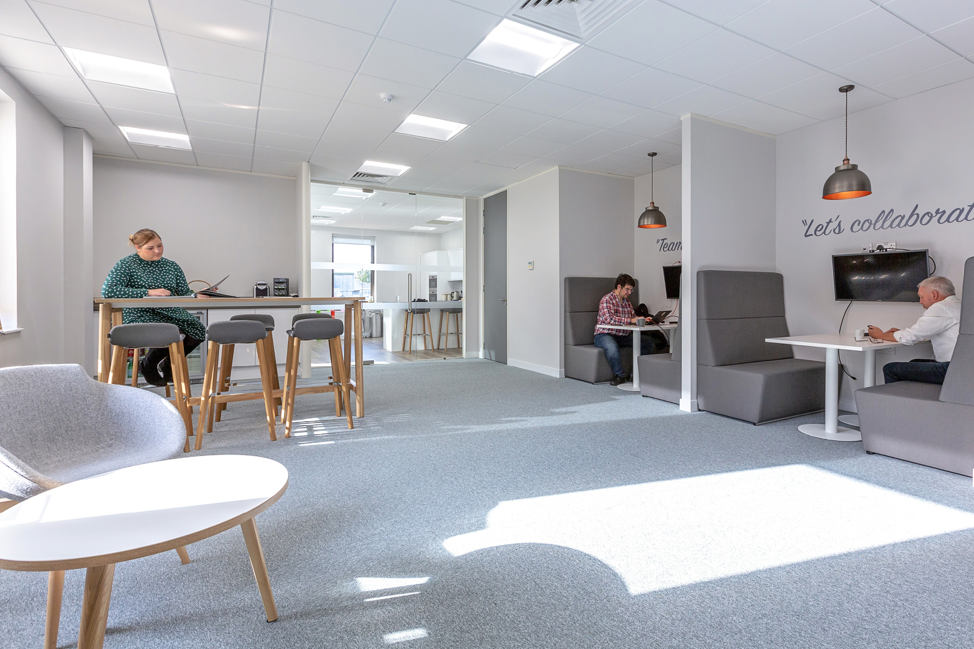 A modern office space with grey carpet and white walls. Three people work at different areas.