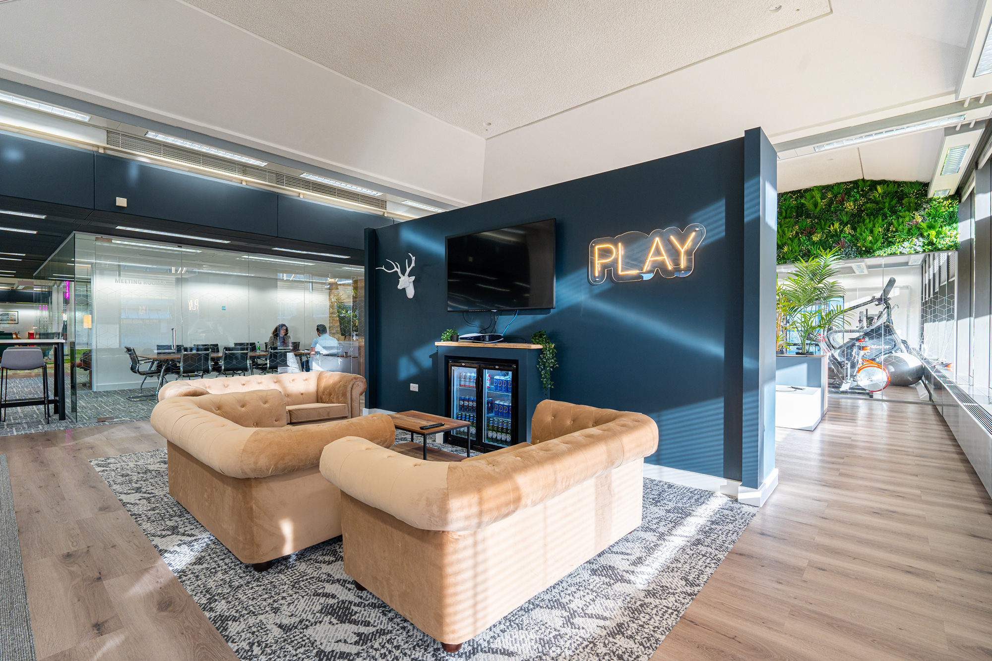 Modern office lounge with two beige sofas around a small table, facing a wall-mounted TV.