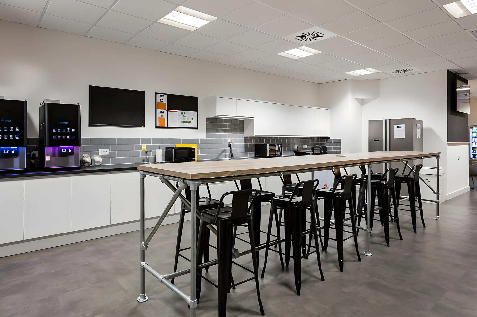 A modern office kitchen features a long wooden table with metal legs surrounded by black stools.