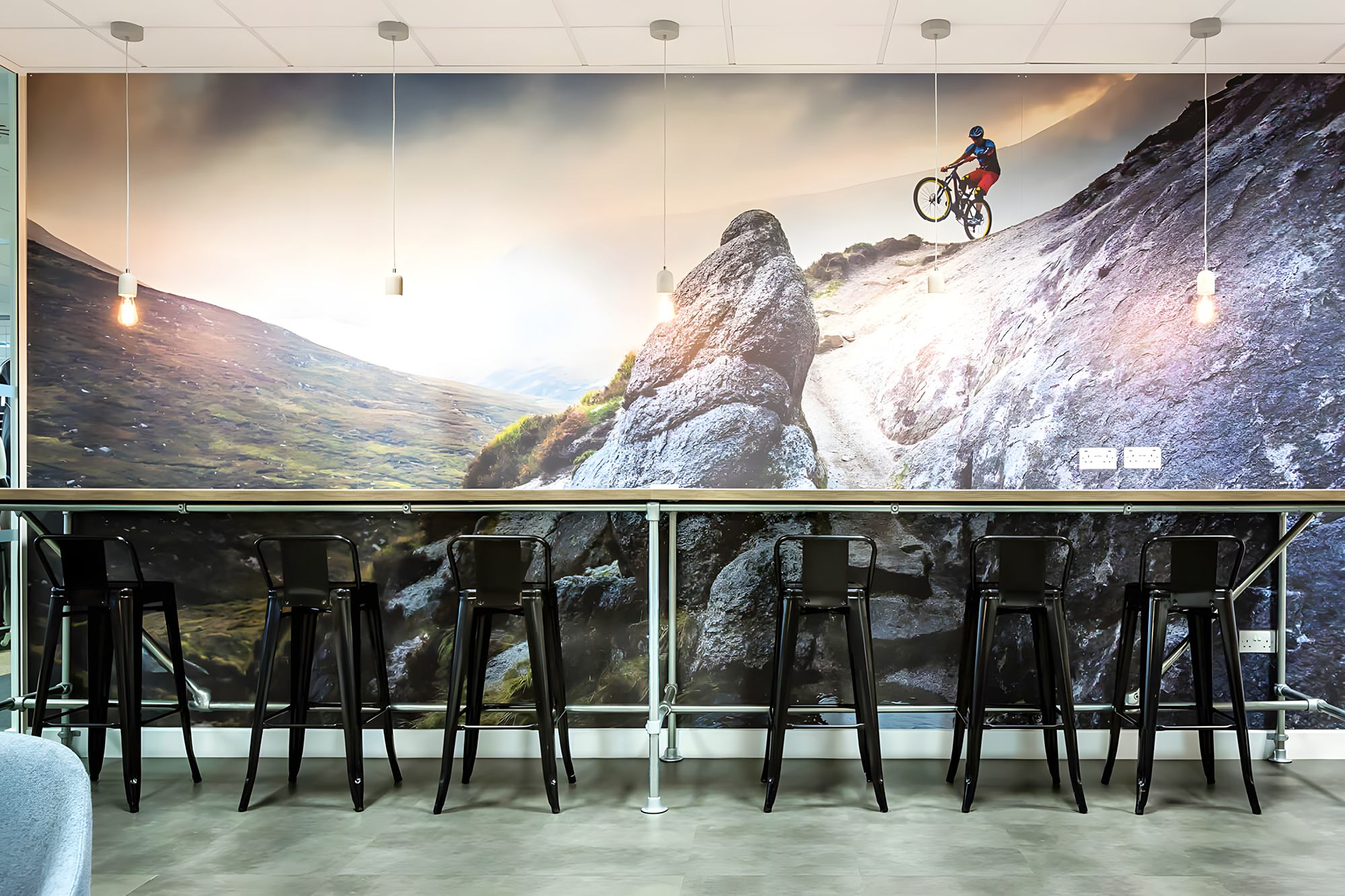 A modern cafe interior with black bar stools and hanging lights. The wall features a large mural of a cyclist.