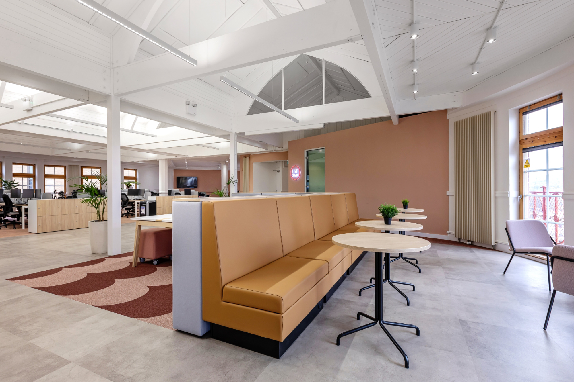 Modern office space with high ceilings and white beams, featuring long tan sofas and round tables.