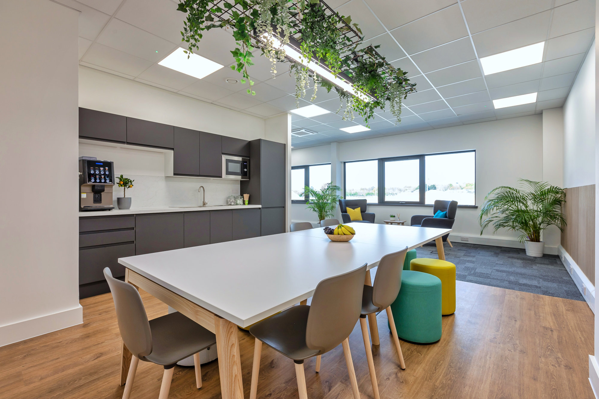 Modern office break room with a long white table, grey chairs, and colourful stools.