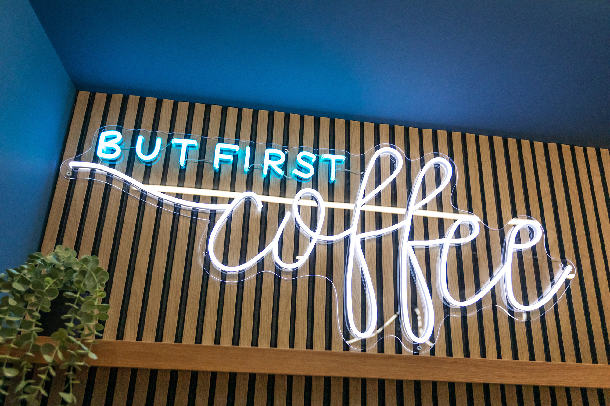 A neon sign on a wooden slat wall reads BUT FIRST coffee. The words BUT FIRST are in blue, and coffee is in white cursive.