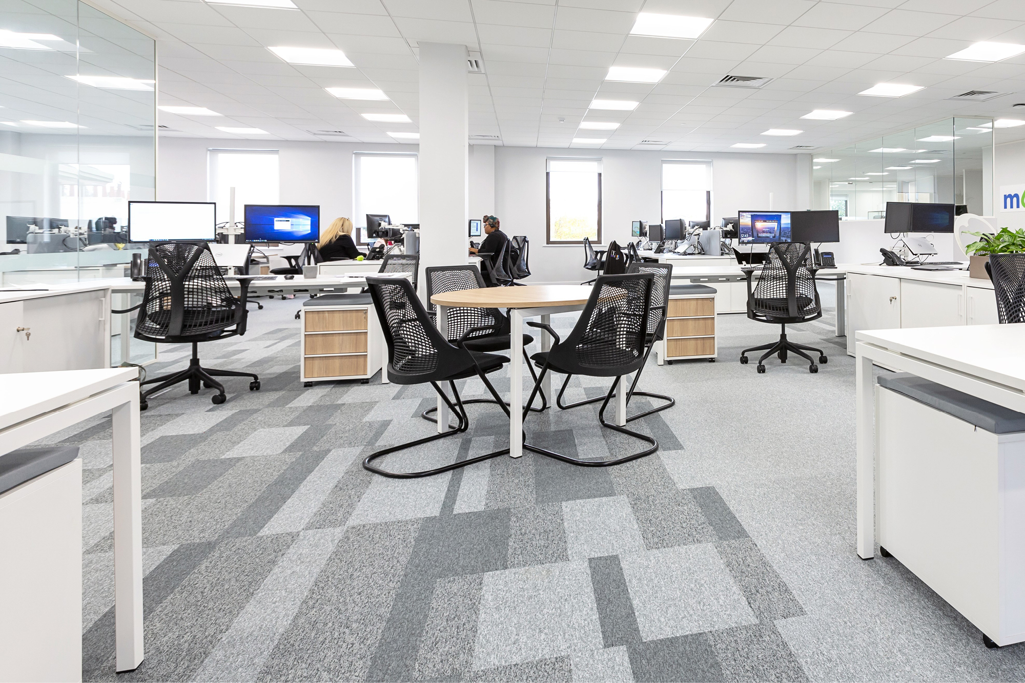 A modern, open-plan office with several workstations featuring black mesh chairs and computer monitors.