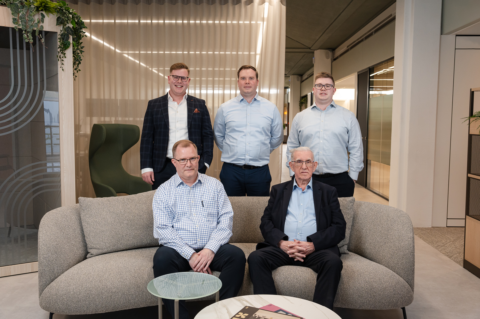 3 men standing and 2 men sitting on beige sofa