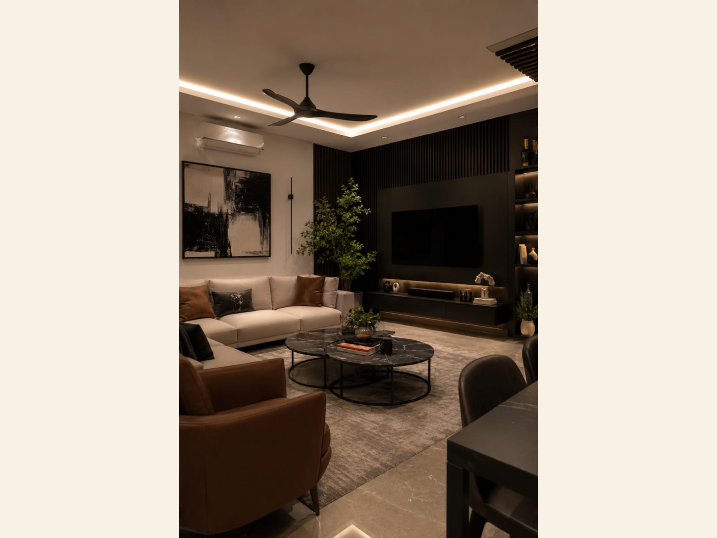 A warm, low-lit living room built around a black fluted TV wall, an ivory sectional, a cognac leather chair and twin round black-marble coffee tables.